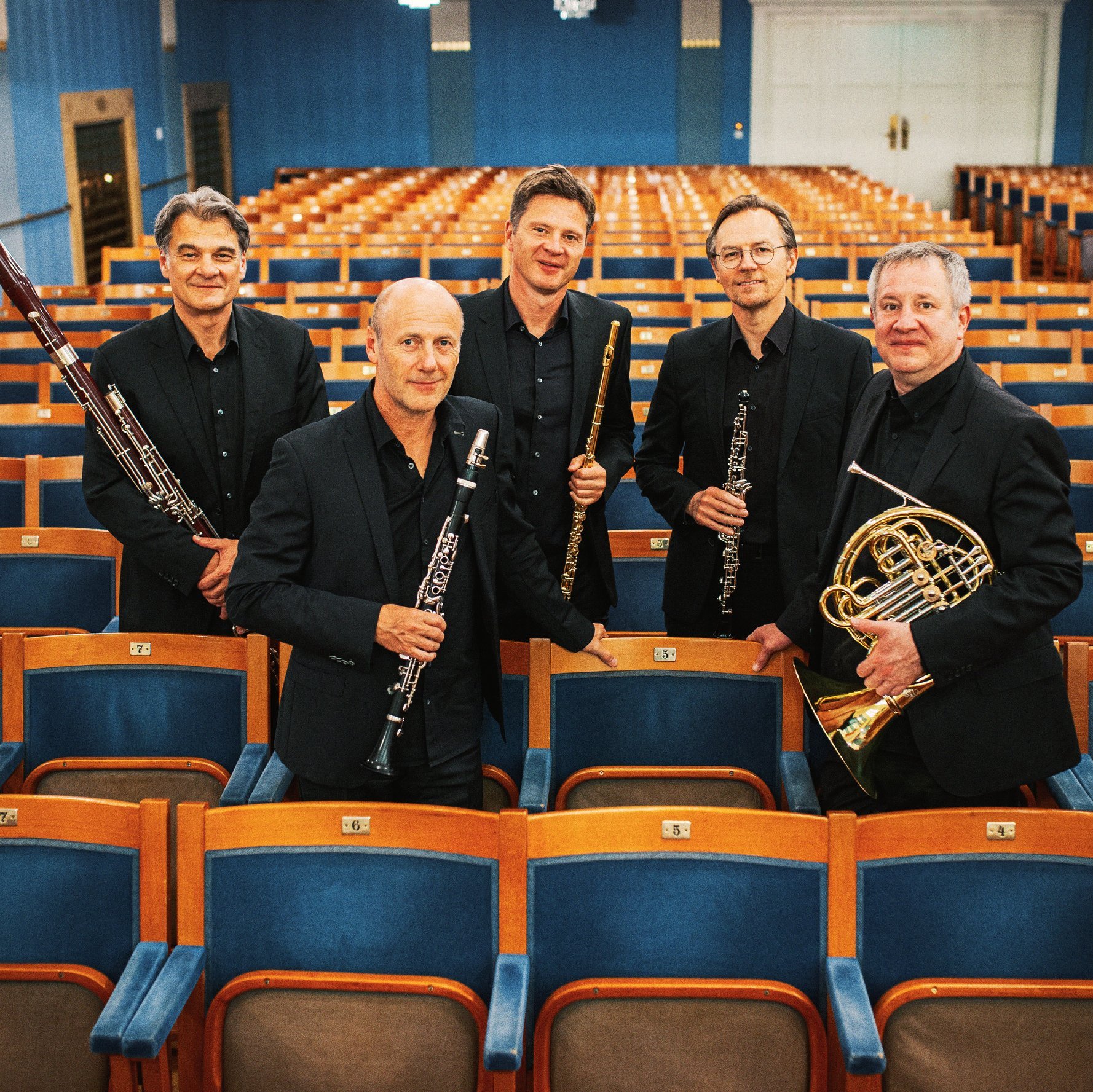 Fünf Männer in schwarzen Anzügen, die Orchesterinstrumente in der Hand halten, stehen inmitten von Reihen leerer Theatersitze und posieren für ein Gruppenfoto in einem hell erleuchteten, eleganten Konzertsaal.