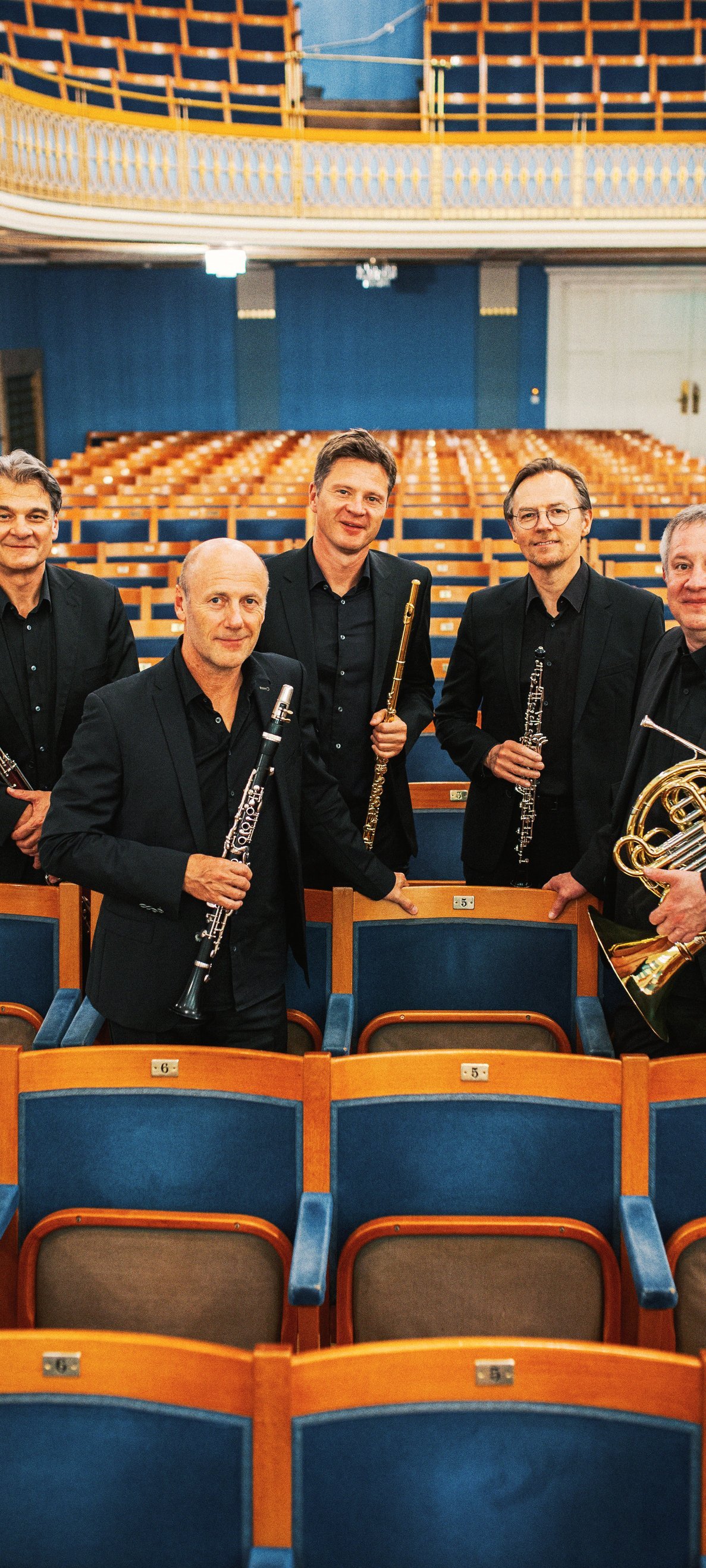 Five men in black suits holding wind instruments stand among empty auditorium seats, posing for a group photo in a bright, elegant concert hall with blue and gold accents.