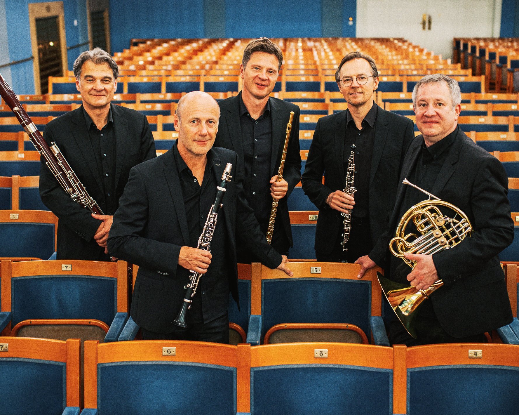 Fünf Männer in schwarzen Anzügen, die Orchesterinstrumente in der Hand halten, stehen inmitten von Reihen leerer Theatersitze und posieren für ein Gruppenfoto in einem hell erleuchteten, eleganten Konzertsaal.