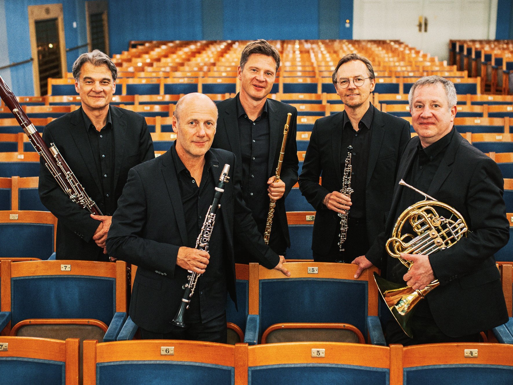 Fünf Männer in schwarzen Anzügen, die Orchesterinstrumente in der Hand halten, stehen inmitten von Reihen leerer Theatersitze und posieren für ein Gruppenfoto in einem hell erleuchteten, eleganten Konzertsaal.