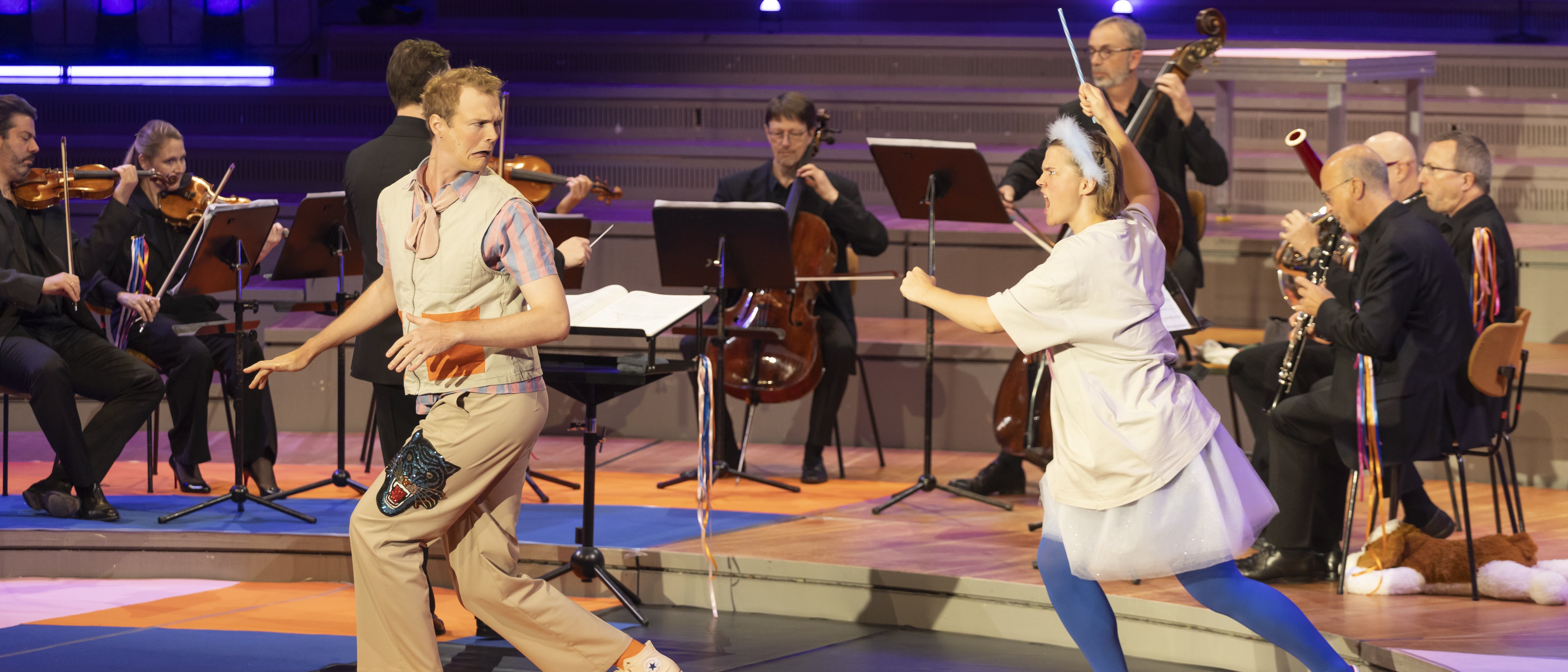 Two performers in imaginative costumes play in front of a chamber orchestra on a colourfully designed stage in the Berlin Philharmonie.
