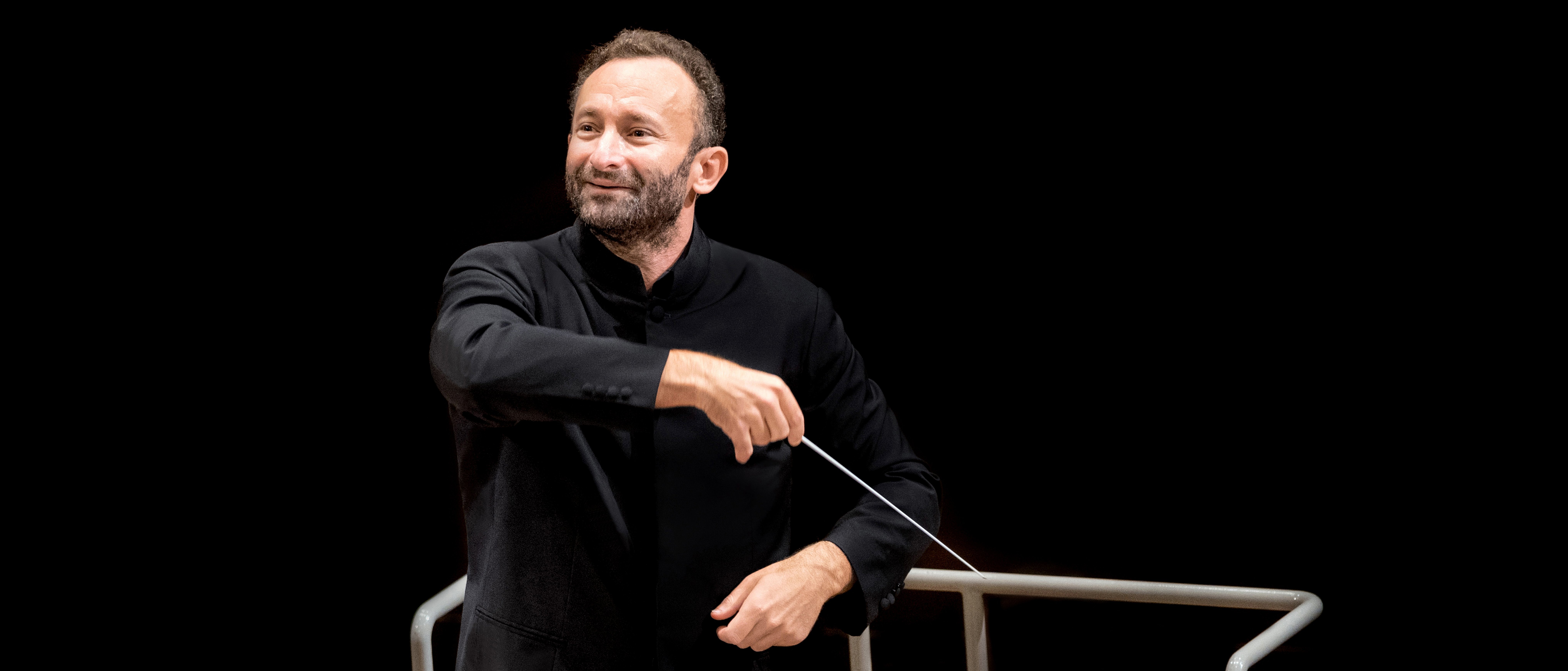 Kirill Petrenko im schwarzen Anzug leitet leidenschaftlich das Orchester. In der Hand hält er einen Taktstock, vor einem dunklen Hintergrund. Vor ihm steht ein Notenständer mit Notenblättern.