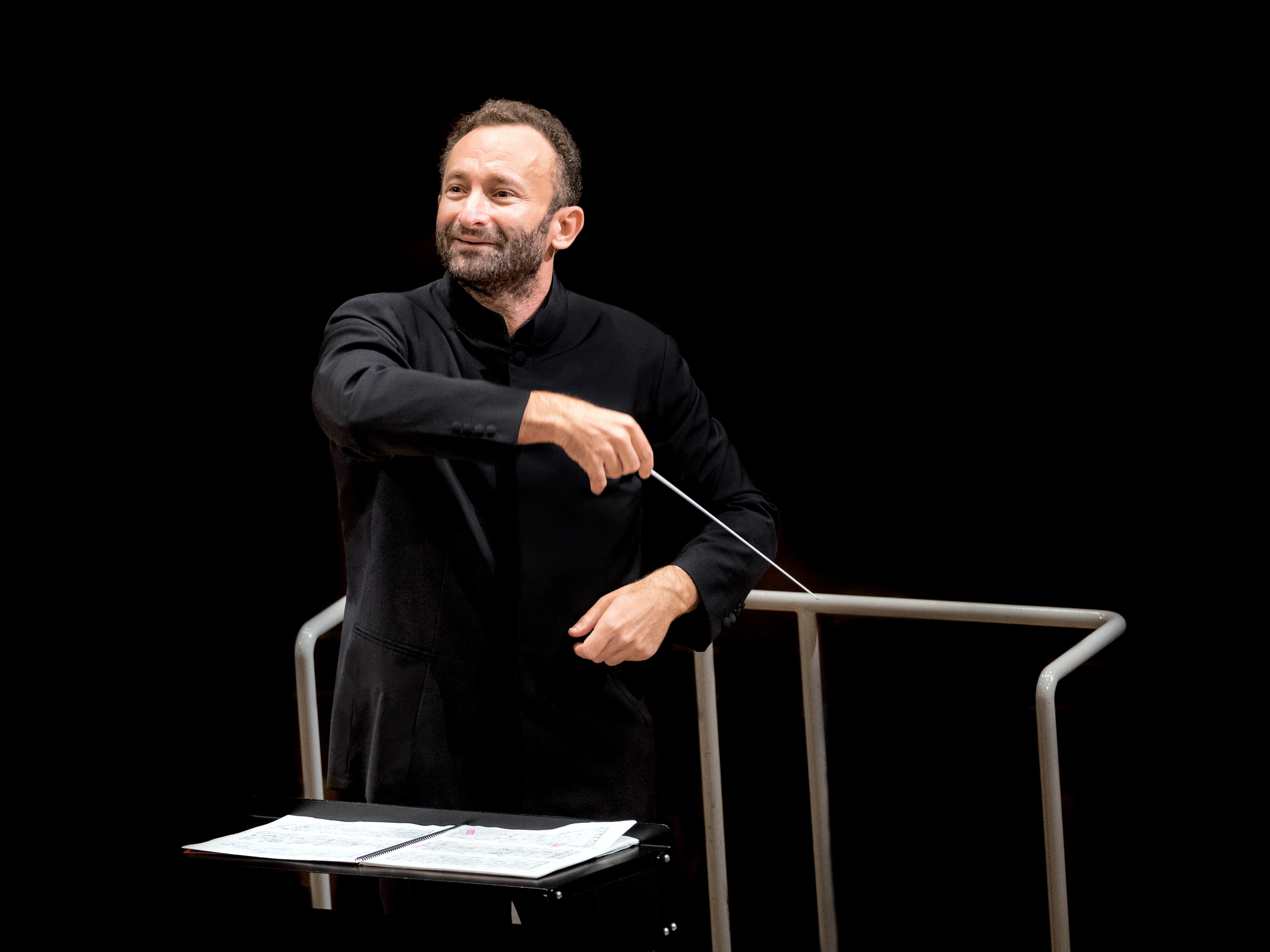 Kirill Petrenko im schwarzen Anzug leitet leidenschaftlich das Orchester. In der Hand hält er einen Taktstock, vor einem dunklen Hintergrund. Vor ihm steht ein Notenständer mit Notenblättern.
