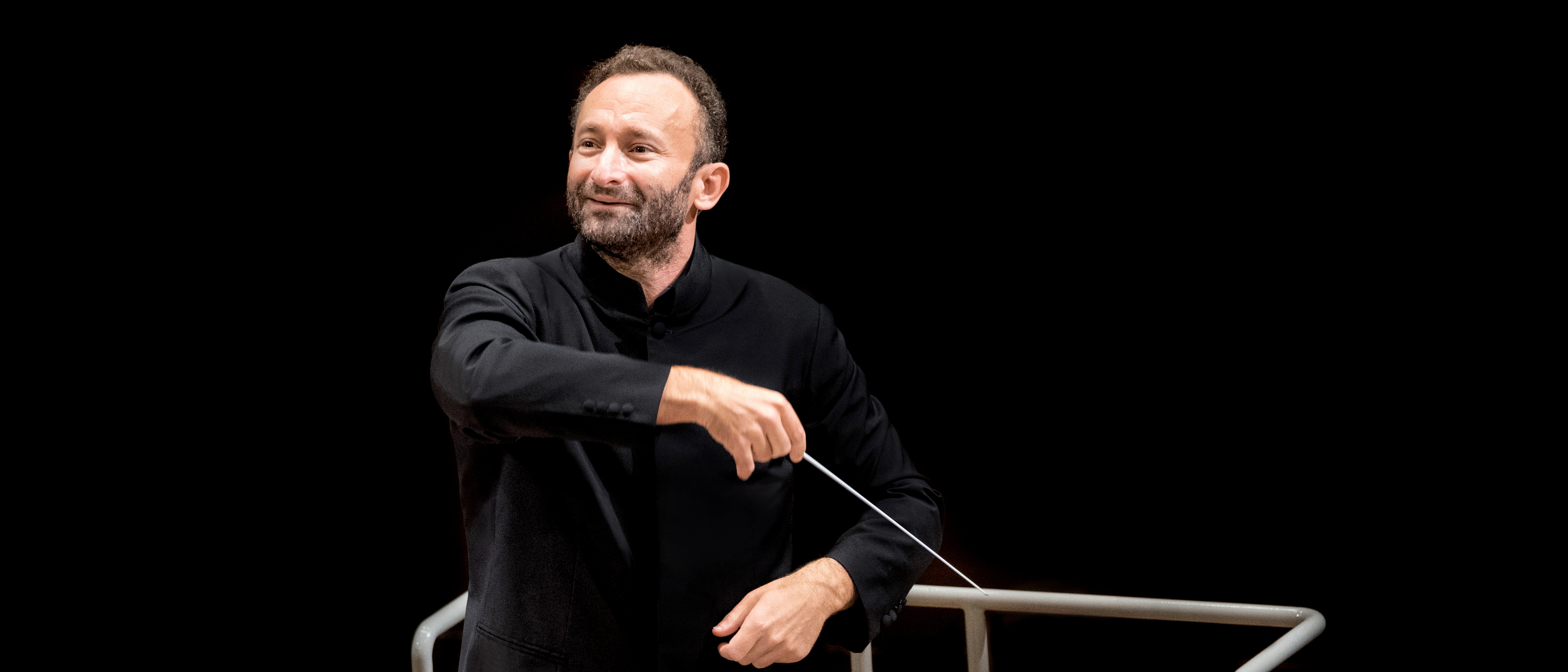 Kirill Petrenko im schwarzen Anzug leitet leidenschaftlich das Orchester. In der Hand hält er einen Taktstock, vor einem dunklen Hintergrund. Vor ihm steht ein Notenständer mit Notenblättern.