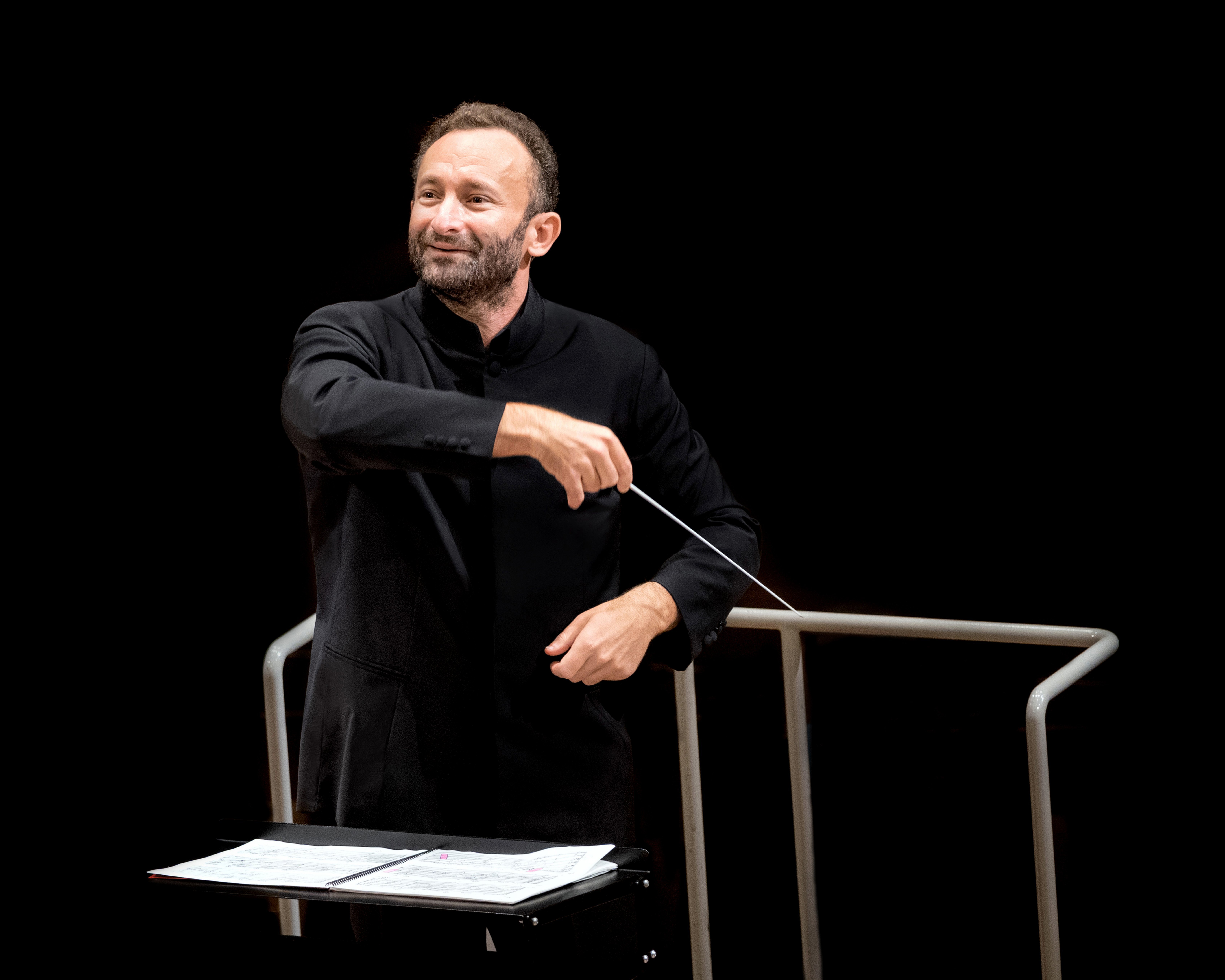 Kirill Petrenko im schwarzen Anzug leitet leidenschaftlich das Orchester. In der Hand hält er einen Taktstock, vor einem dunklen Hintergrund. Vor ihm steht ein Notenständer mit Notenblättern.