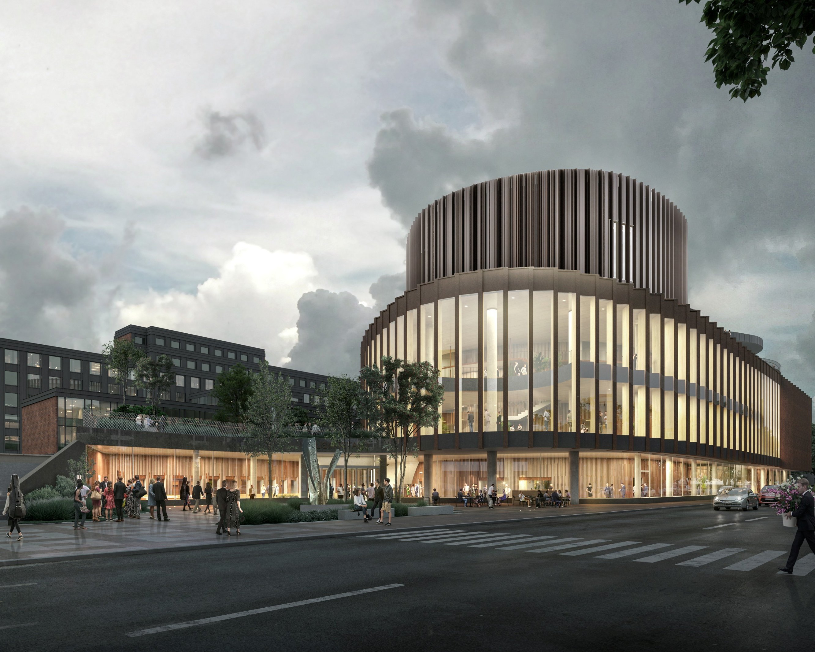 A modern, multi-story building with curved architecture and large glass windows is illuminated at dusk. People walk nearby, and cars drive on the street in front of the building under a cloudy sky.