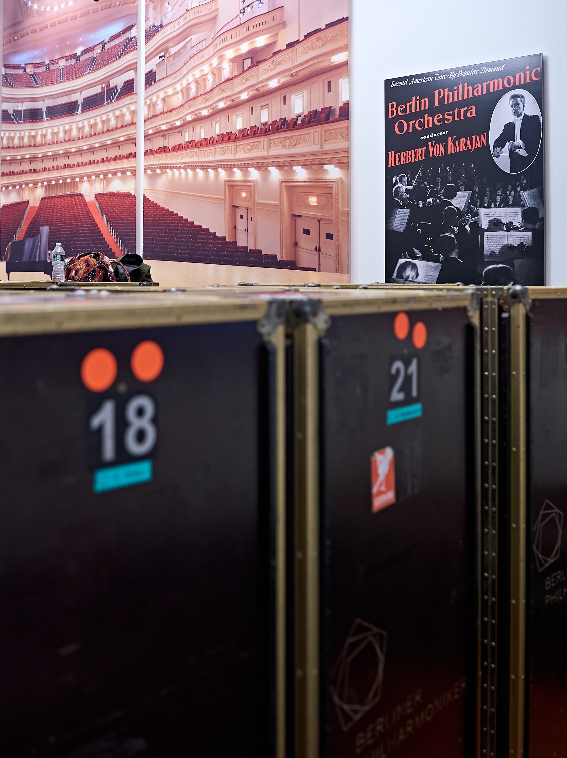 Ein großes Wandbild, das einen leeren Konzertsaal hinter mehreren schwarzen Instrumentenkoffern zeigt, von denen einer mit der Nummer 18 beschriftet ist, sowie ein gerahmtes Konzertplakat der Berliner Philharmoniker.