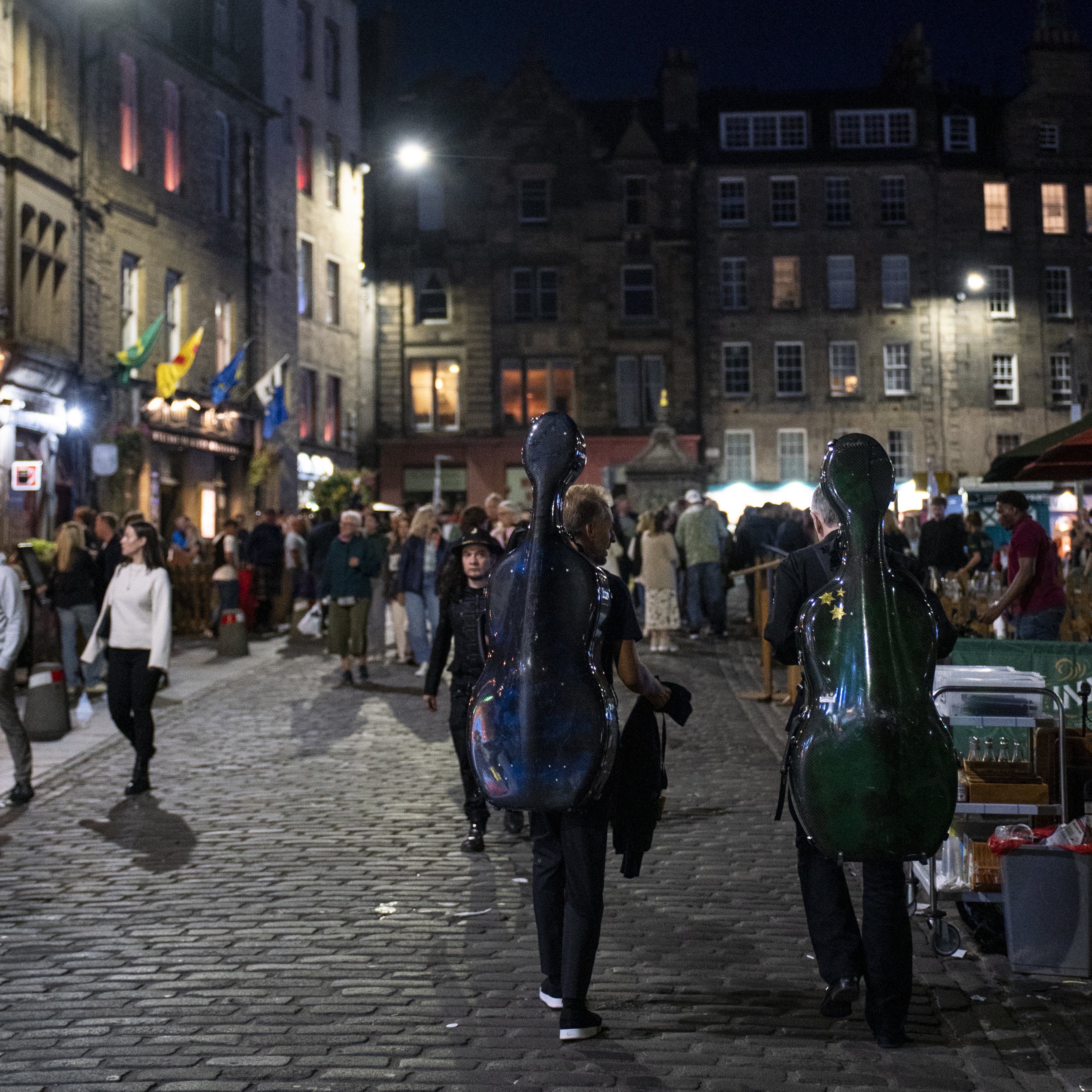 Two people carrying cello cases walk down a cobblestone street at night, surrounded by a crowd and illuminated by streetlights, with historic buildings in the background.