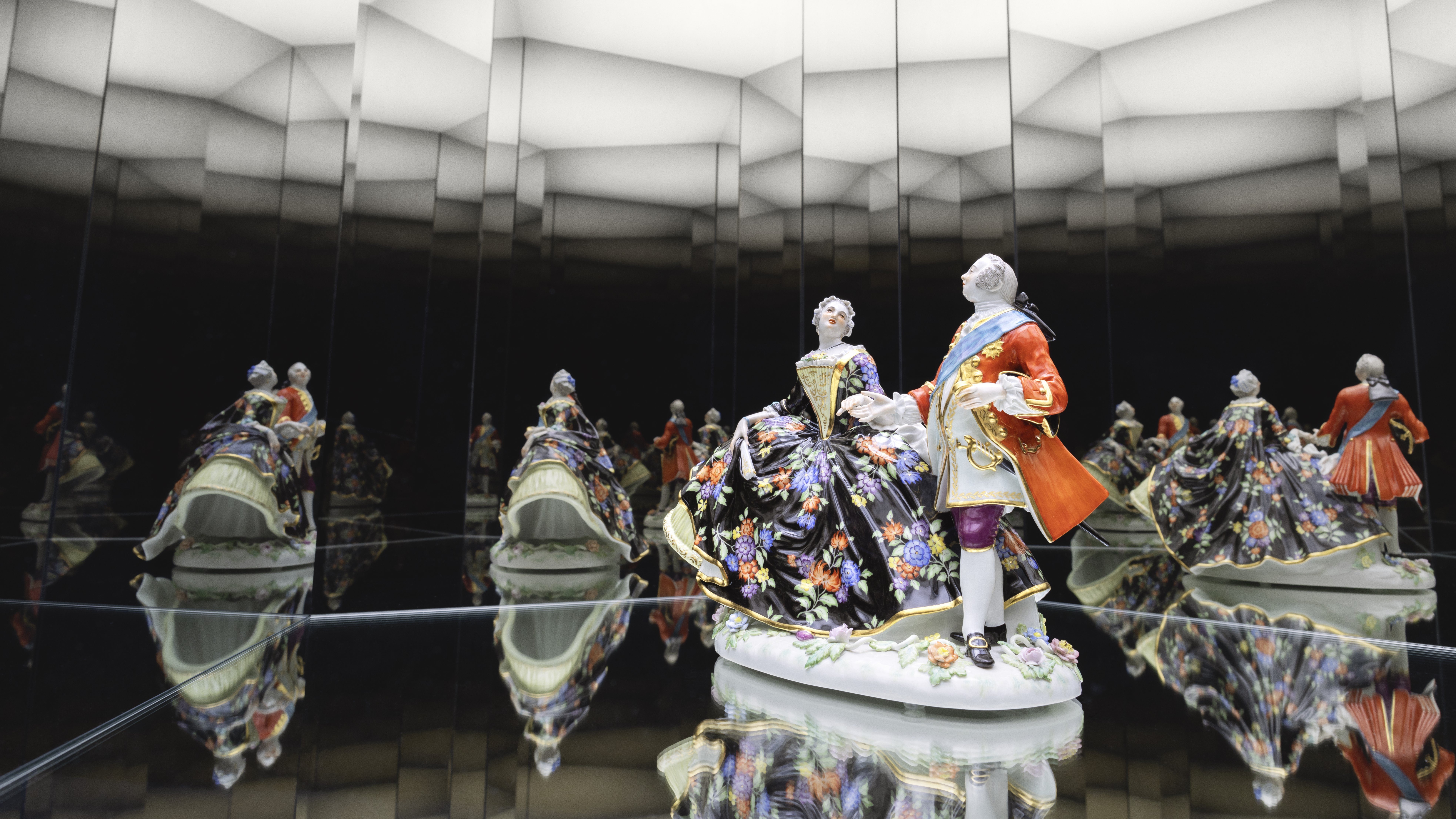 Eine Porzellanfigur eines Mannes und einer Frau in kunstvoller, historischer Kleidung steht auf einer Spiegelfläche, die von den umliegenden Spiegeln in einem geometrischen, beleuchteten Raum mehrfach reflektiert wird.