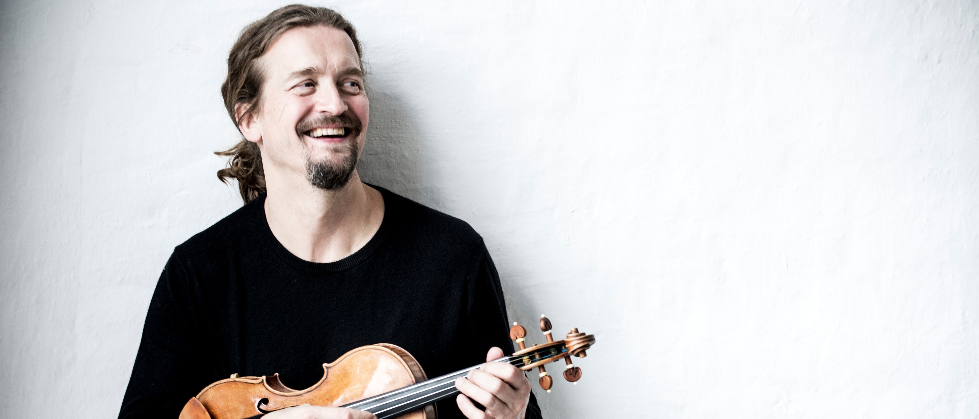 Christian Teztlaff mit langen Haaren in einem Pferdeschwanz, einem schwarzen Hemd und hellen Jeans steht vor einer weißen Wand und hält eine Geige und einen Bogen in den Händen.