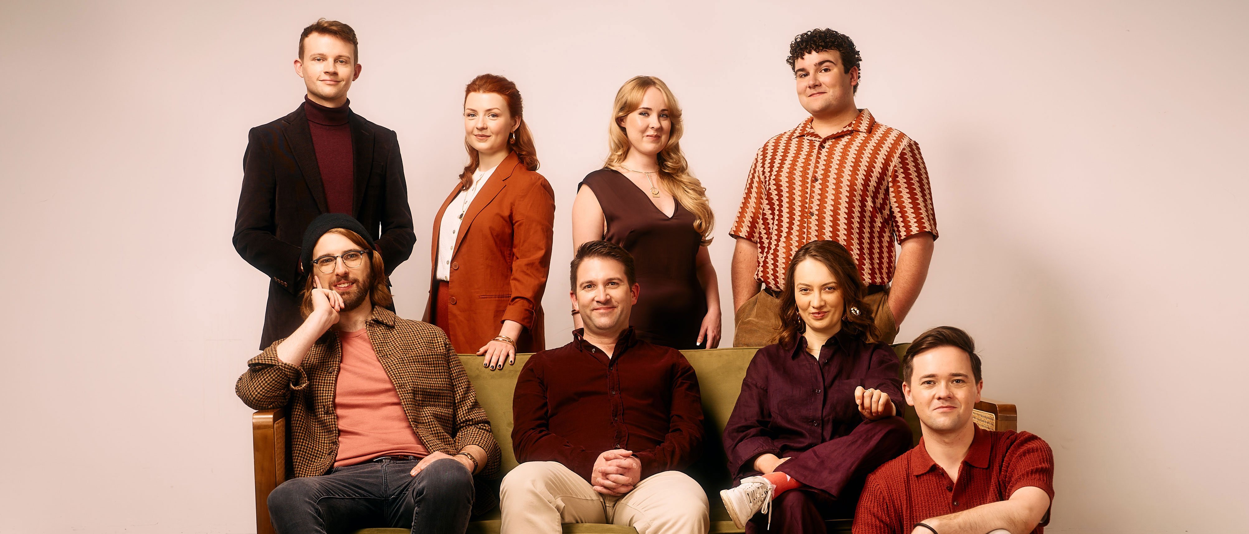 Acht Erwachsene posieren für ein Gruppenfoto in einem Studio. Drei stehen hinter einer grünen Couch, fünf sitzen oder lehnen sich an sie. Sie tragen legere und halboffizielle Kleidung. Der Hintergrund ist einfarbig, und der Boden ist aus Holz.