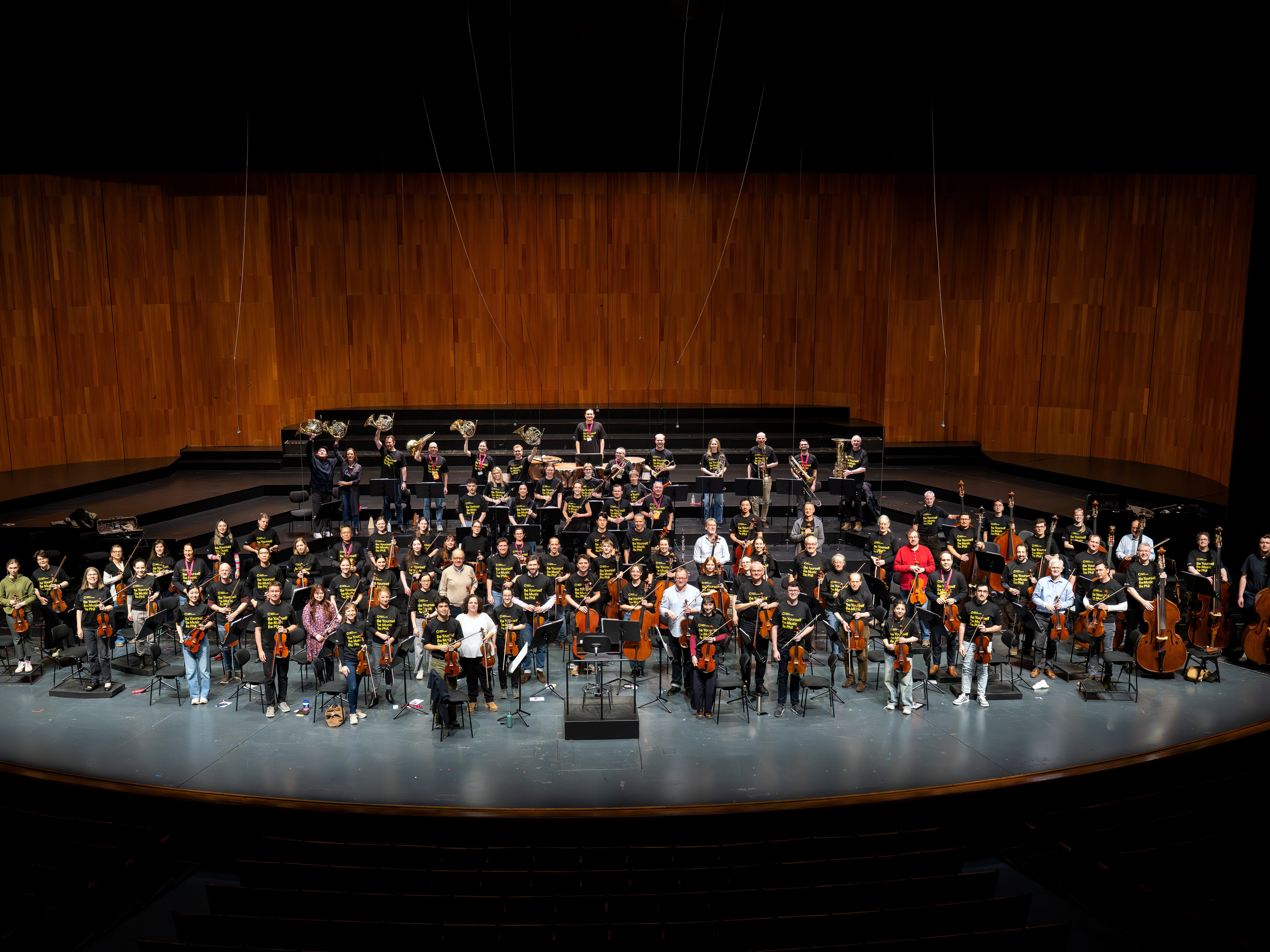 Ein großes Symphonieorchester mit Musikern, die verschiedene Instrumente halten, ist auf einer breiten Bühne in einem modernen Konzertsaal mit Holzwänden angeordnet.