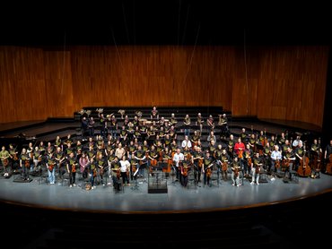 Ein großes Symphonieorchester mit Musikern, die verschiedene Instrumente halten, ist auf einer breiten Bühne in einem modernen Konzertsaal mit Holzwänden angeordnet.