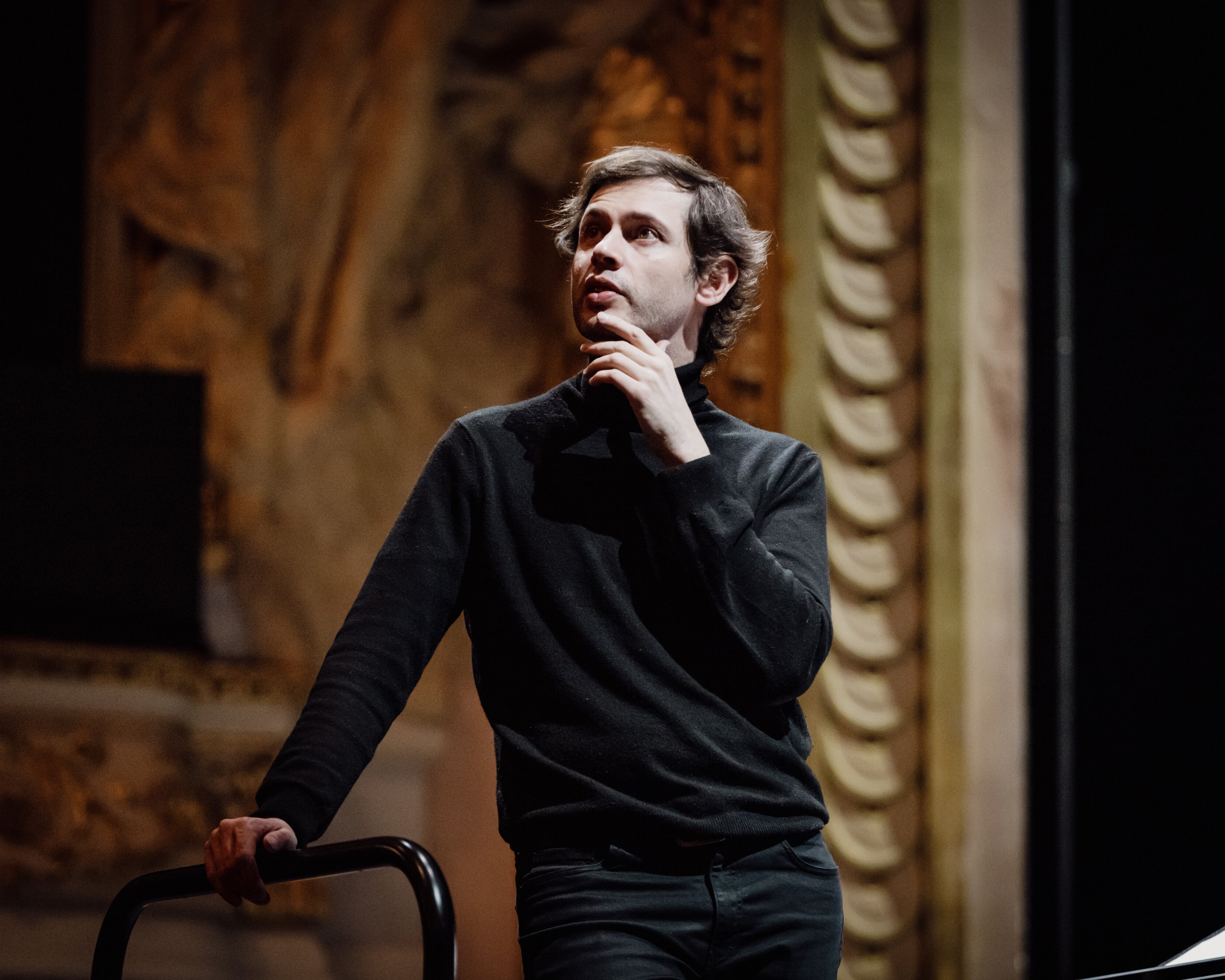 Maxime Pascal in einem schwarzen Pullover steht in einem Haus, stützt sich mit einer Hand auf ein Geländer und berührt nachdenklich sein Kinn, während im Hintergrund kunstvolle architektonische Details zu sehen sind.