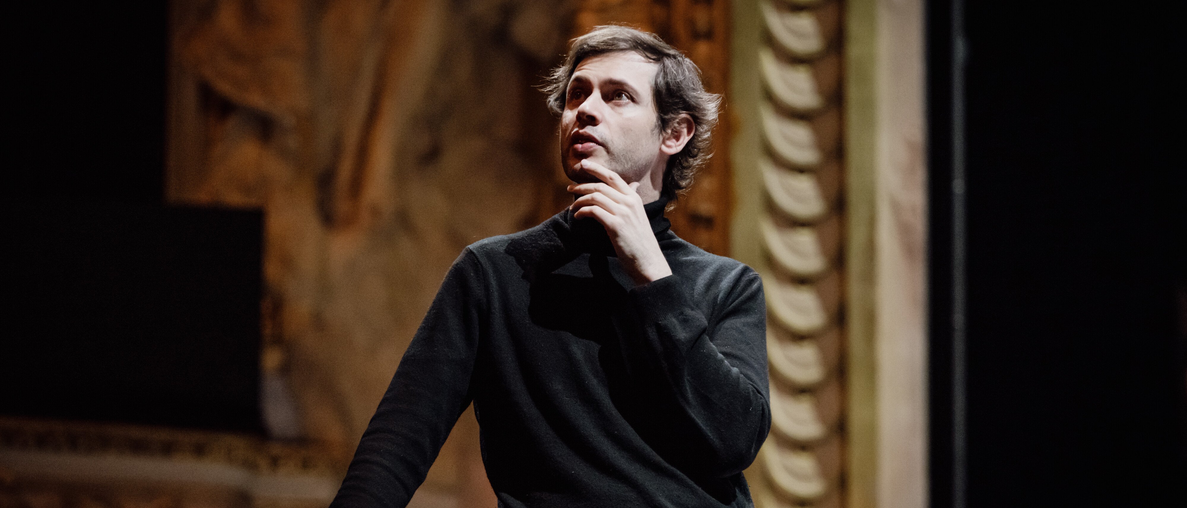 Maxime Pascal in einem schwarzen Pullover steht in einem Haus, stützt sich mit einer Hand auf ein Geländer und berührt nachdenklich sein Kinn, während im Hintergrund kunstvolle architektonische Details zu sehen sind.