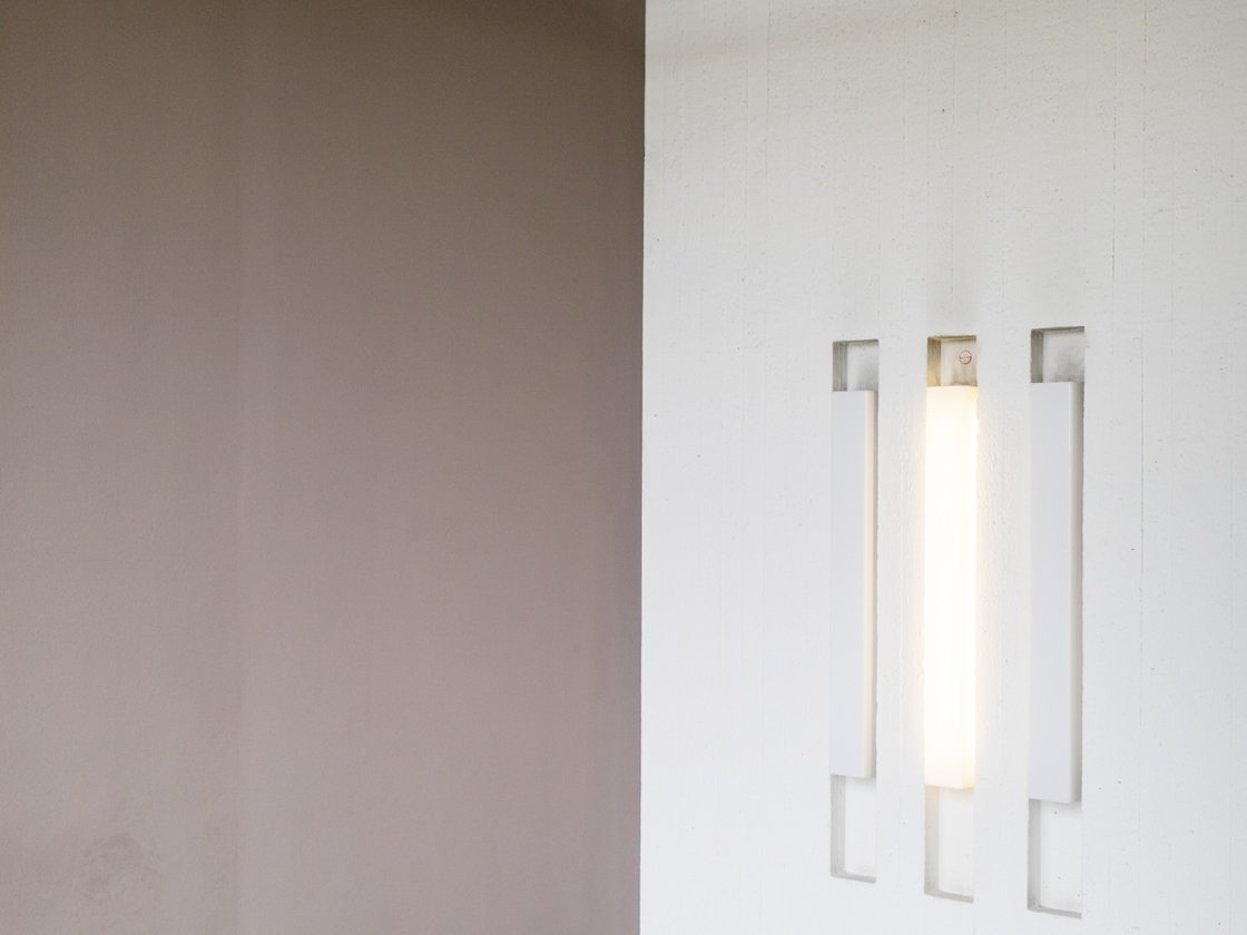 Minimalistisches Interieur mit einer mauvefarbenen Wand, die auf eine weiße Wand trifft, in der sich drei vertikale, rechteckige Aussparungen befinden; in der mittleren Aussparung befindet sich eine leuchtende Leuchte. Boden und Decke sind schlicht und hell gehalten.