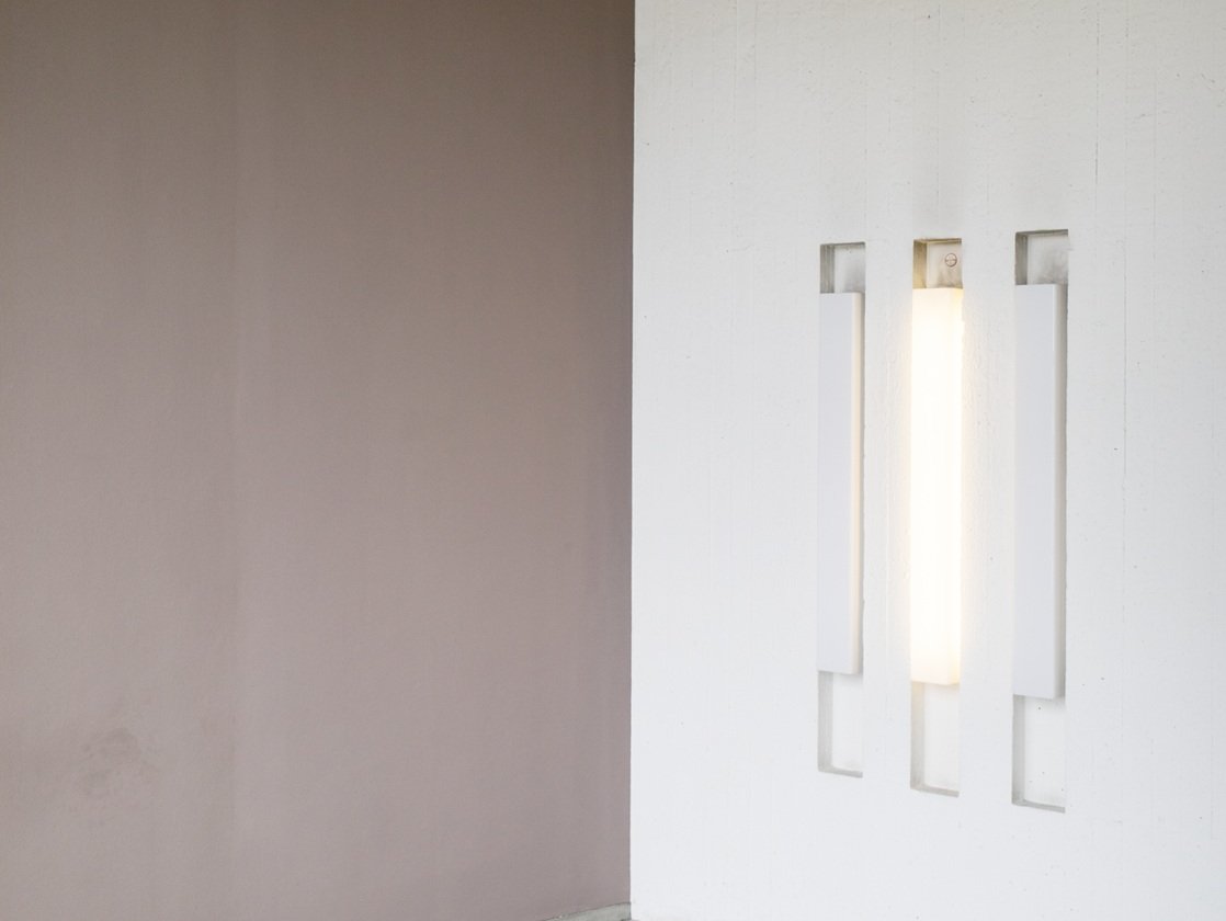 Minimalistisches Interieur mit einer mauvefarbenen Wand, die auf eine weiße Wand trifft, in der sich drei vertikale, rechteckige Aussparungen befinden; in der mittleren Aussparung befindet sich eine leuchtende Leuchte. Boden und Decke sind schlicht und hell gehalten.