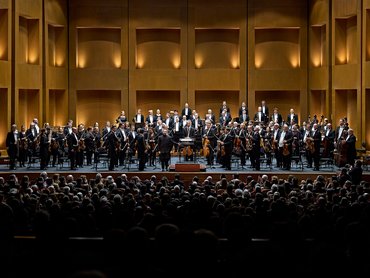 Ein vollbesetztes Orchester spielt auf einer Holzbühne vor einem sitzenden Publikum in einem Konzertsaal.