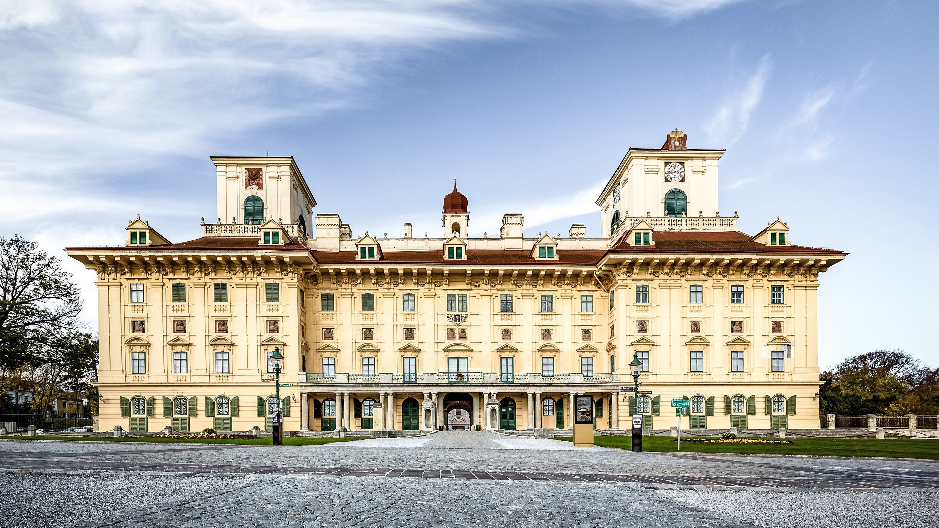 Das Bild zeigt ein prächtiges, kunstvolles Gebäude mit einer kunstvollen Fassade, symmetrischem Design und einem zentralen, gewölbten Eingang. Es verfügt über drei Türme mit Uhren und steht vor einem strahlend blauen Himmel.