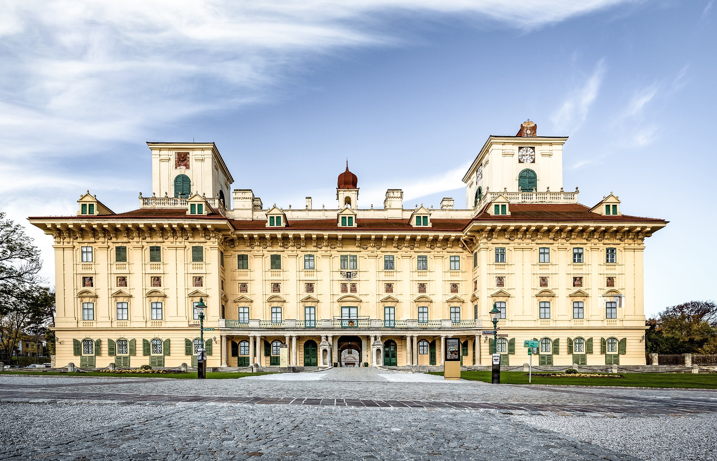 The picture shows a magnificent, ornate building with an elaborate façade, symmetrical design and a central, arched entrance. It has three towers with clocks and stands against a bright blue sky.
