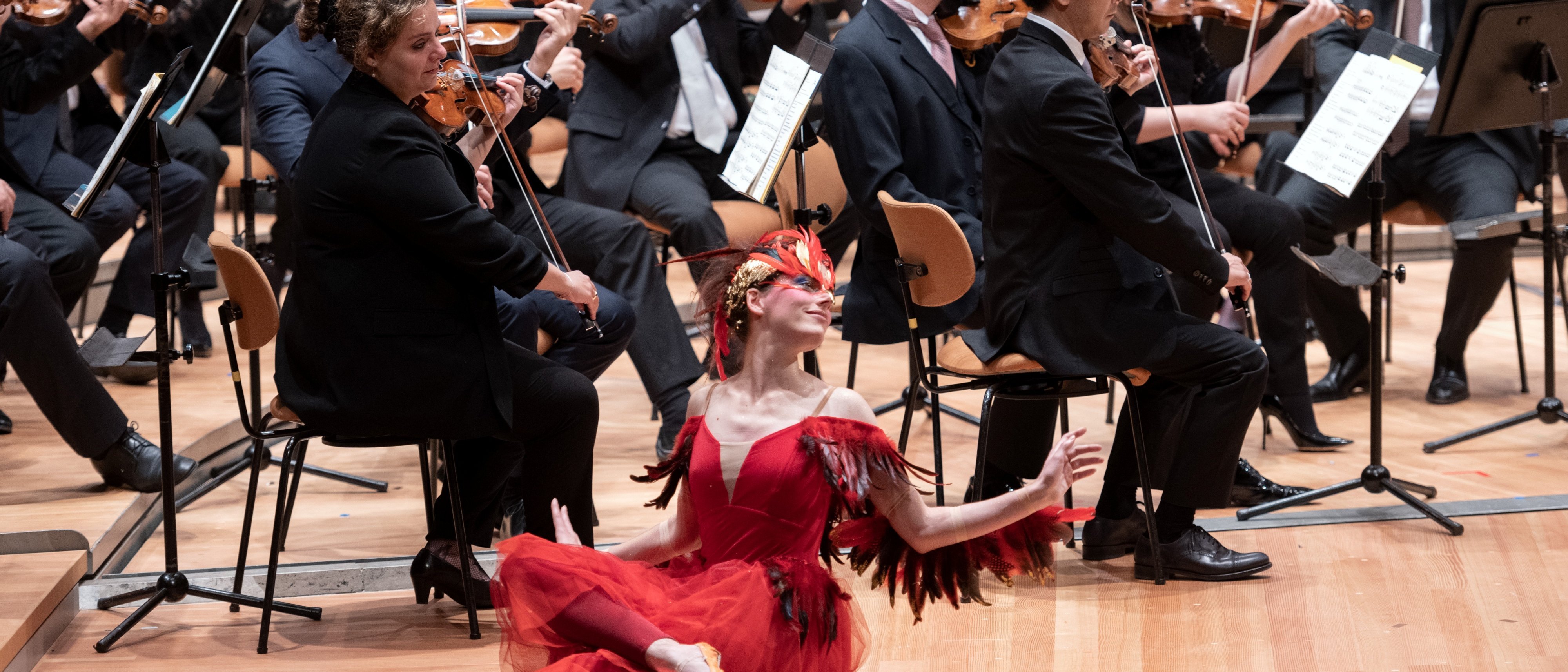Eine Ballerina in einem roten Federkostüm sitzt auf der Bühne und posiert anmutig vor einem vollen Orchester, dessen Musiker in formeller schwarzer Kleidung Streichinstrumente spielen.