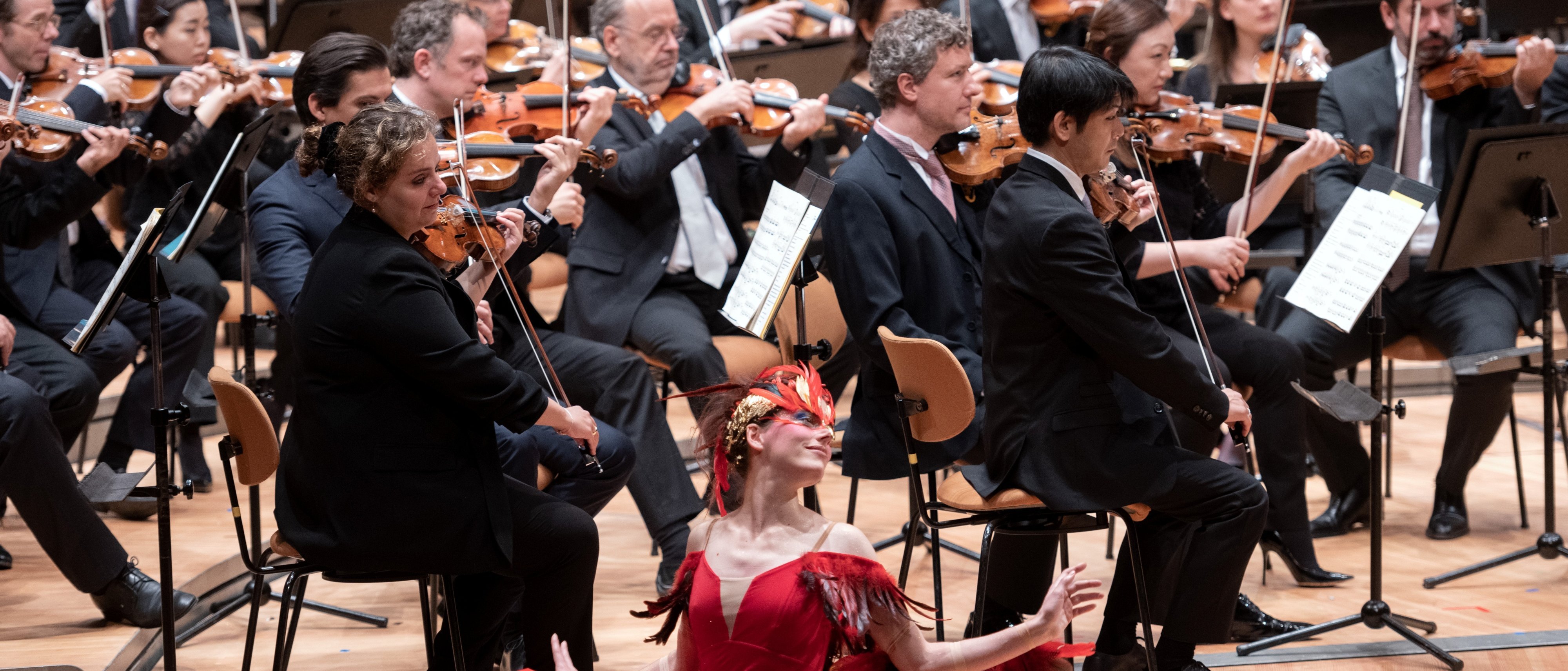 Eine Ballerina in einem roten Federkostüm sitzt auf der Bühne und posiert anmutig vor einem vollen Orchester, dessen Musiker in formeller schwarzer Kleidung Streichinstrumente spielen.