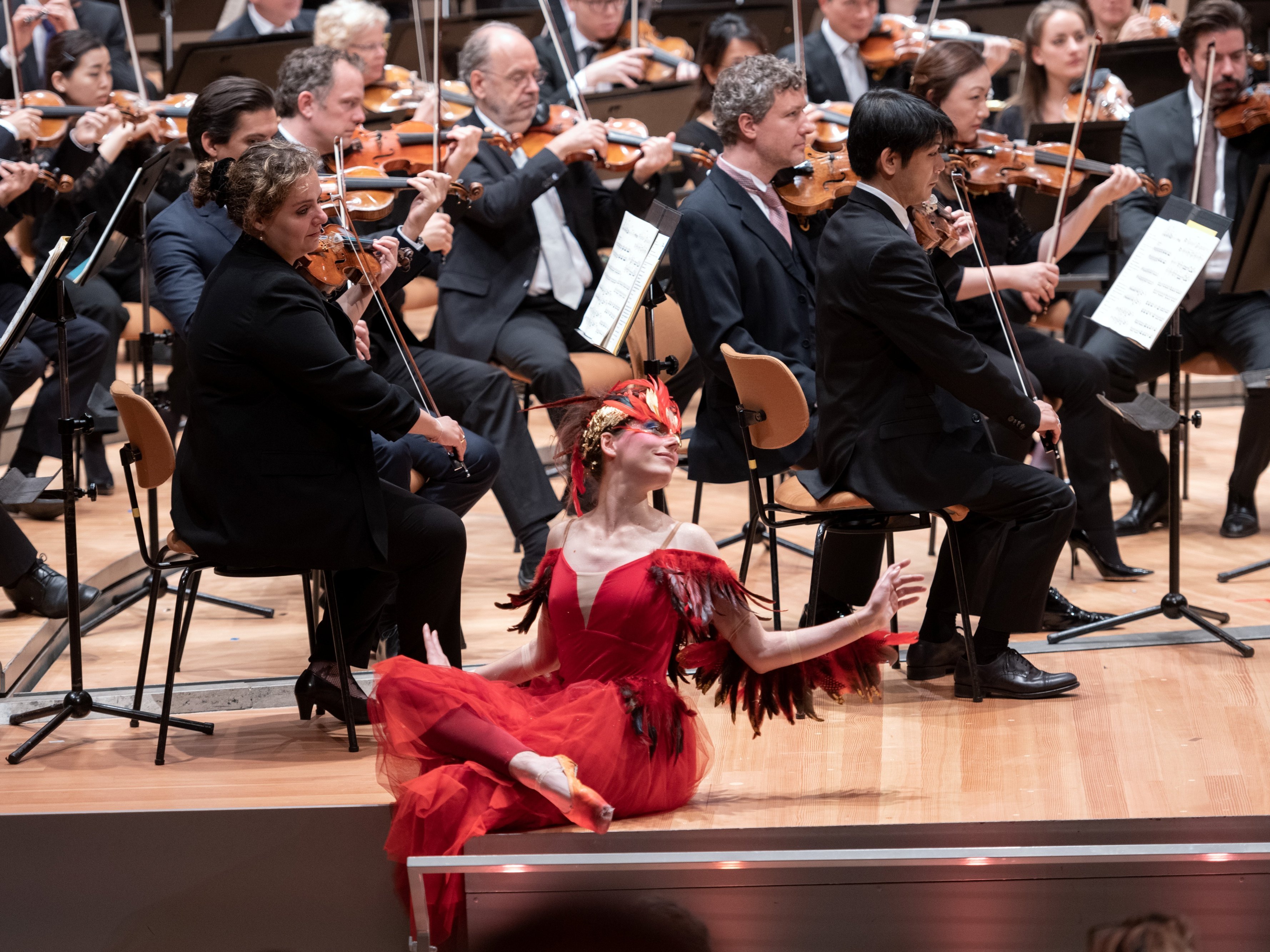 Eine Ballerina in einem roten Federkostüm sitzt auf der Bühne und posiert anmutig vor einem vollen Orchester, dessen Musiker in formeller schwarzer Kleidung Streichinstrumente spielen.