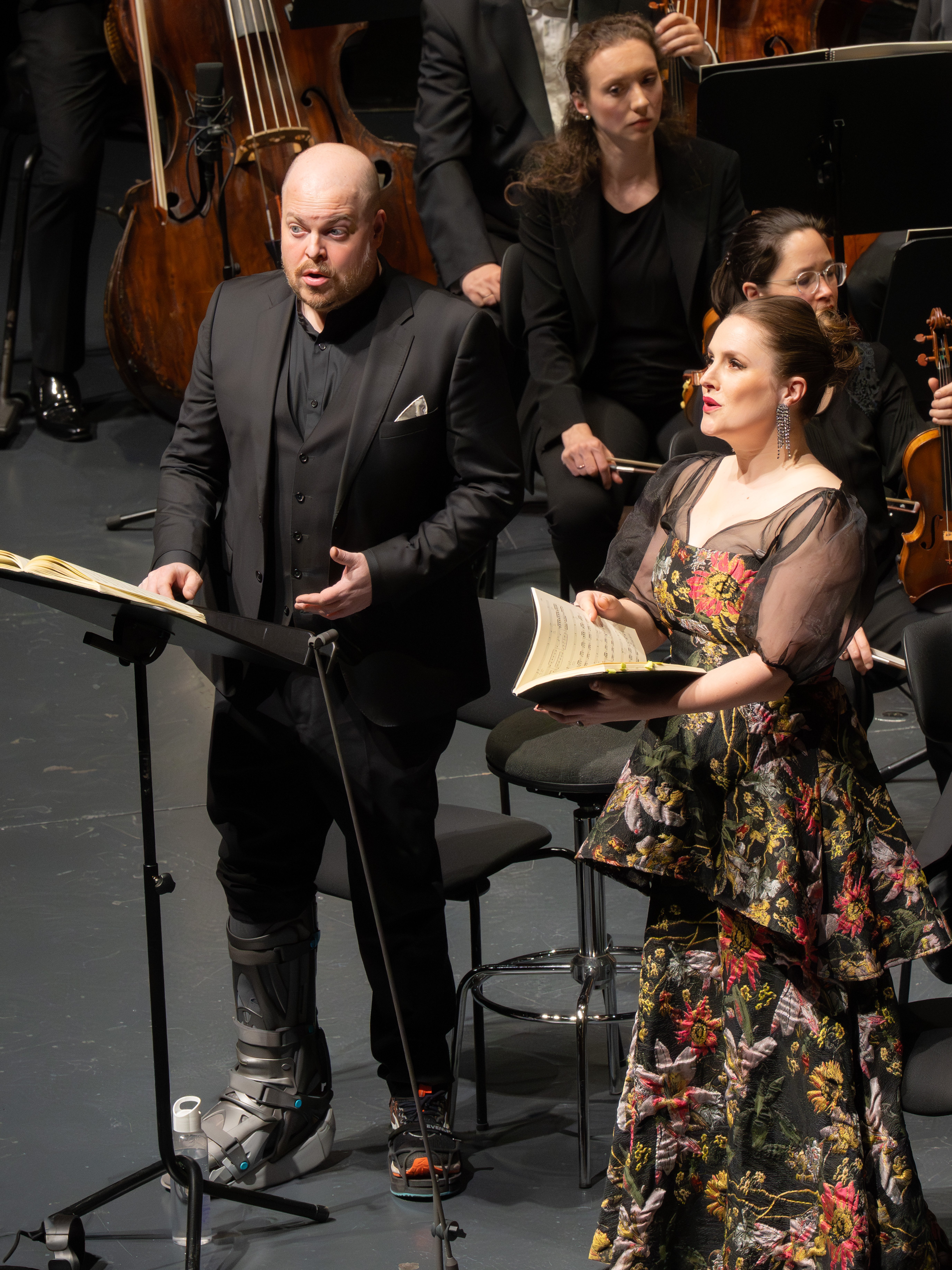 Ein Mann in Anzug und eine Frau in einem geblümten Kleid halten Notenblätter in der Hand und singen auf der Bühne, während ein Orchester hinter ihnen steht.