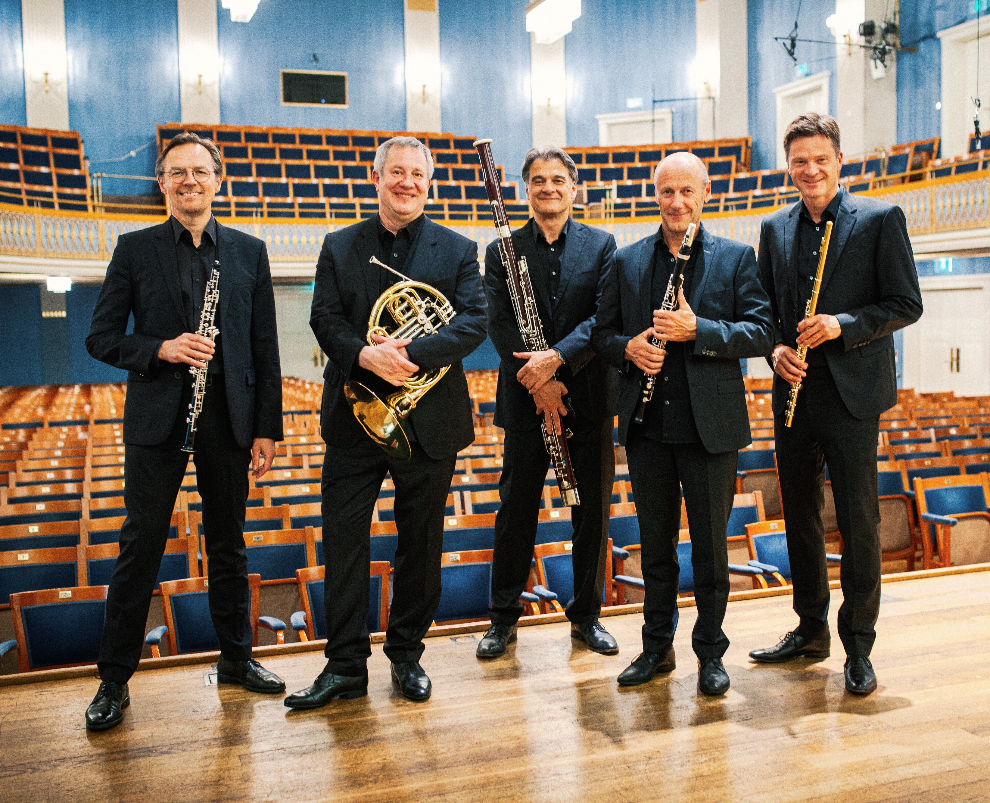 Fünf Musiker in schwarzen Anzügen stehen auf der Bühne eines Konzertsaals und halten Blasinstrumente in der Hand. Hinter ihnen befinden sich leere Reihen von Holzsitzen und blaue Wände mit weißer Umrandung.