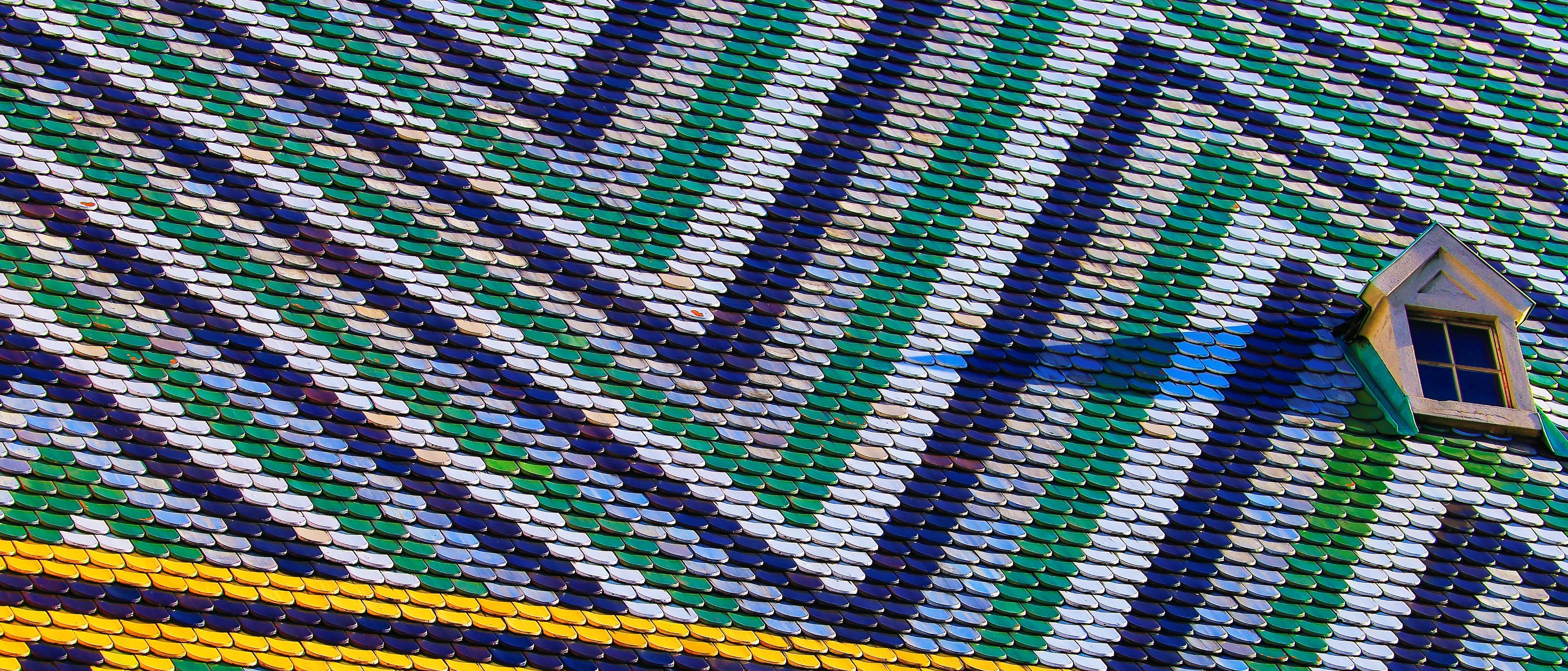 Bunte Nahaufnahme des gemusterten Daches des Stephansdoms in Wien mit Zickzack- und Diagonalmustern aus grünen, gelben, weißen und dunkelblauen Ziegeln, mit zwei kleinen Dachfenstern.