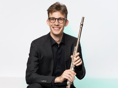A smiling young man in a black suit and glasses holds a silver flute upright in both hands against a plain white background with a faint green tint.