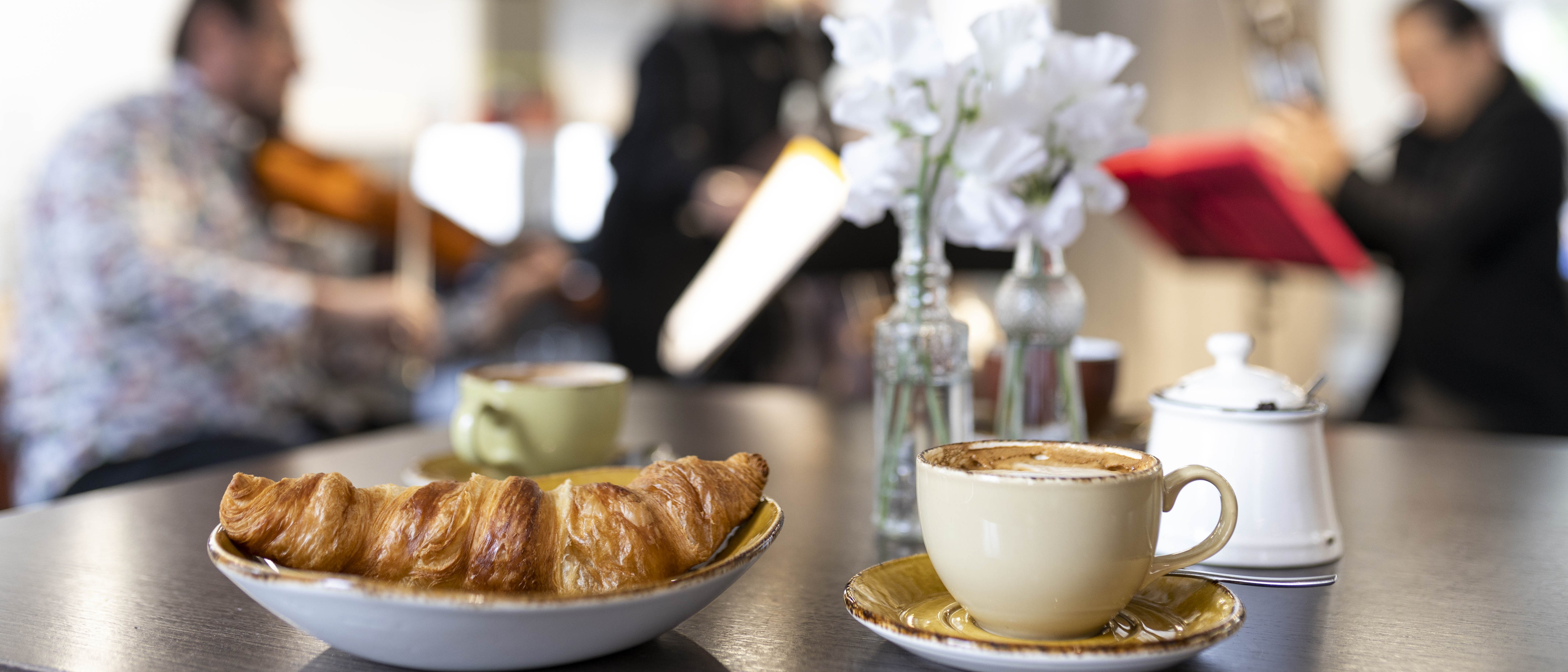 Croissant and coffee on a table, musicians play in the background