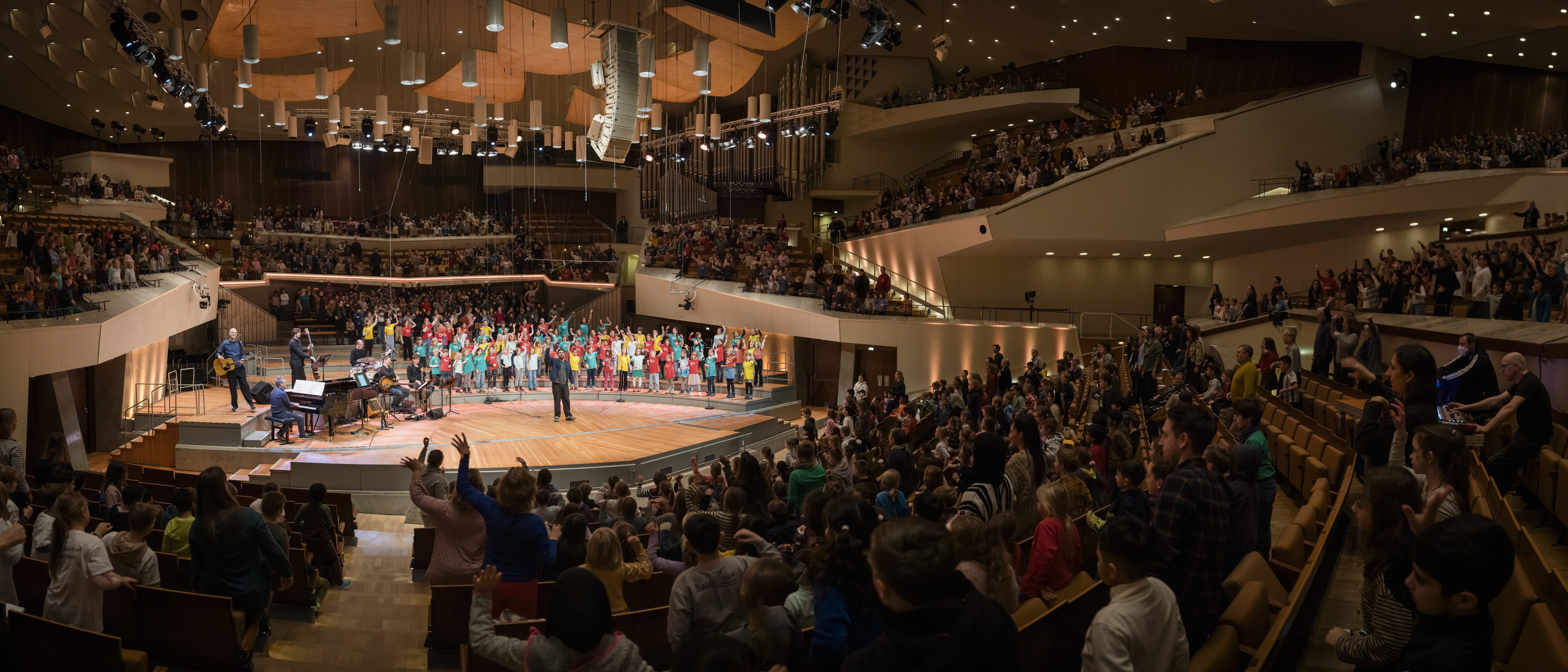 Ein großer Chor in farbenfrohen Kostümen tritt auf der Bühne eines voll besetzten Konzertsaals auf. Die umliegenden Sitzplätze sind mit Zuschauern besetzt, einige stehen. Der Saal ist in warmes Licht getaucht.