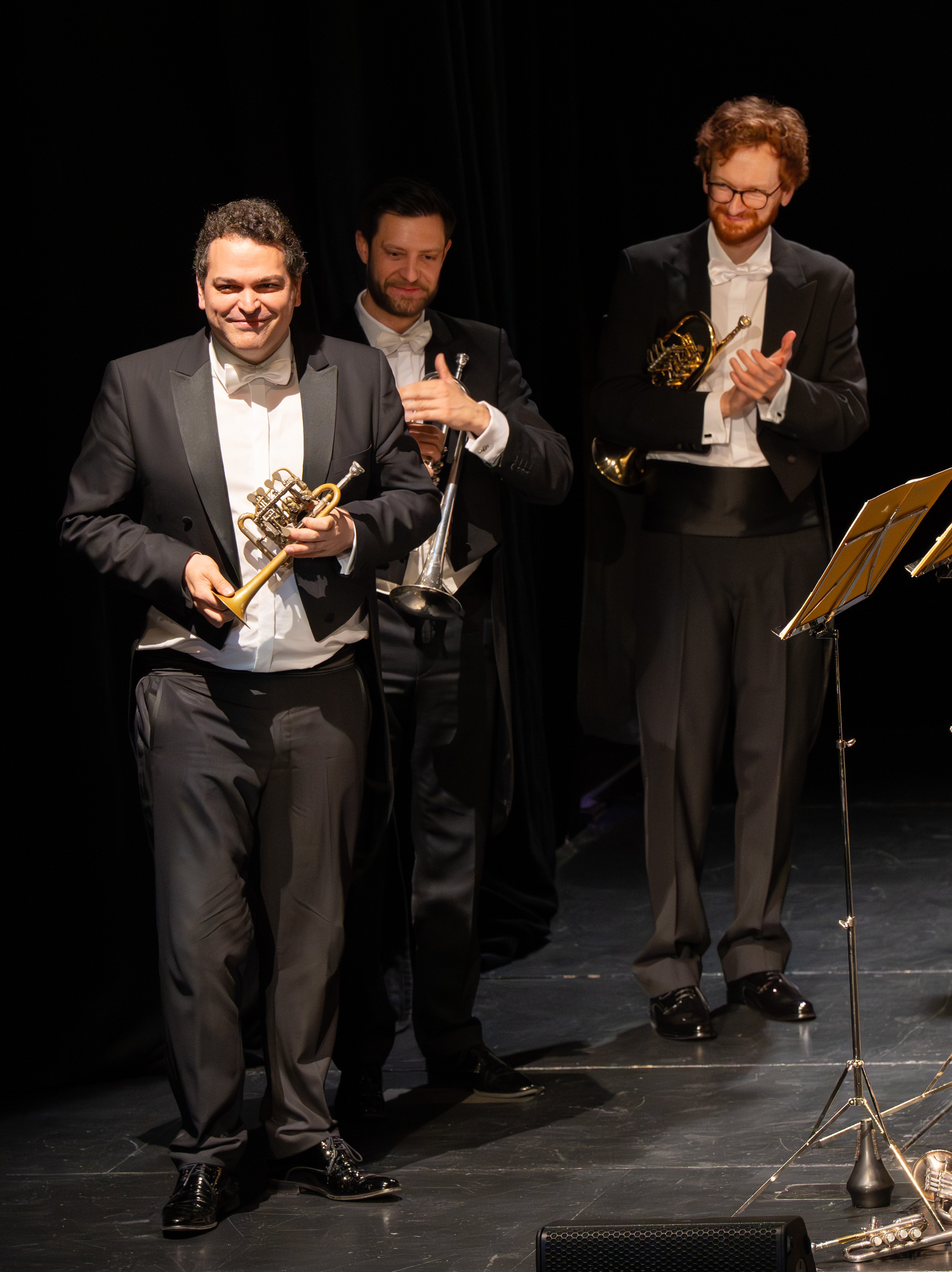 Drei formell gekleidete Männer stehen auf einer Bühne mit Musikinstrumenten; der vordere Mann hält ein Waldhorn, die beiden hinteren halten und klatschen mit Blechblasinstrumenten neben einem Notenständer.