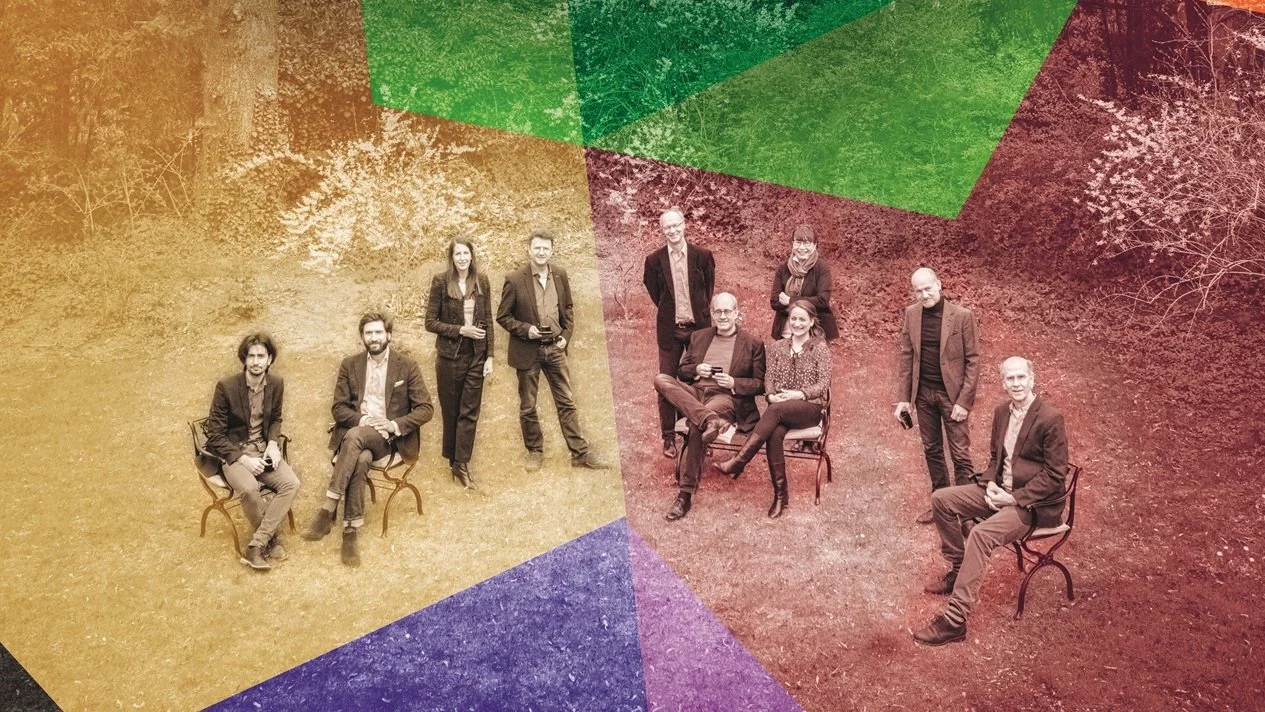 Acht Personen sitzen auf Stühlen auf einem Rasen in einem Garten, mit großen Bäumen und einem Haus im Hintergrund. Transparente, bunte geometrische Formen überlagern die Szene und verleihen ihr einen modernen, künstlerischen Effekt.