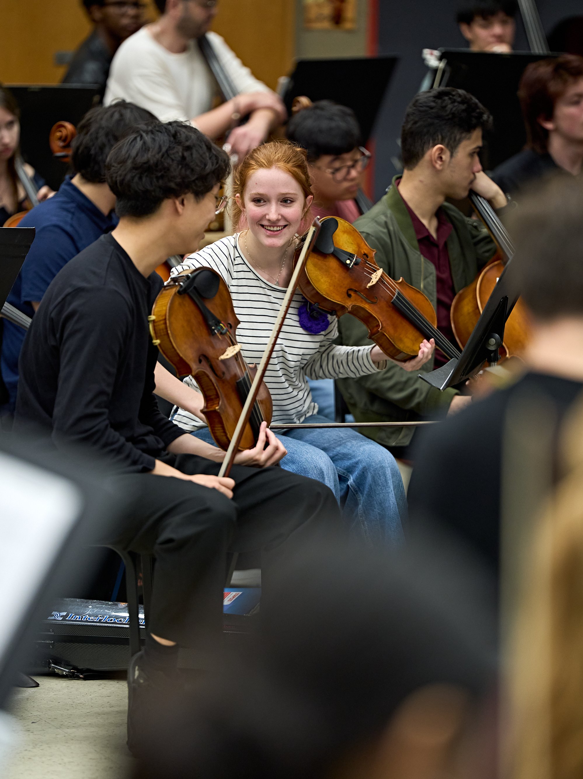 A young woman smiling while playing the violin in an orchestra, seated next to a fellow violinist.