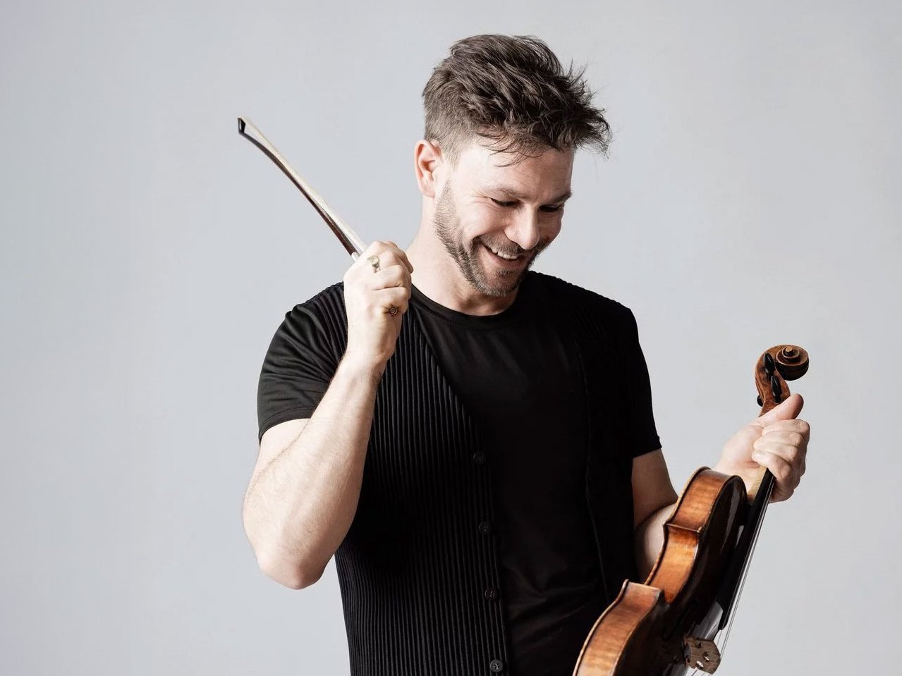 Amihai Grosz lächelnd in einem schwarzen Hemd hält seine Bratsche in einer Hand und hebt spielerisch einen Bogen in der anderen vor einem schlichten hellgrauen Hintergrund.