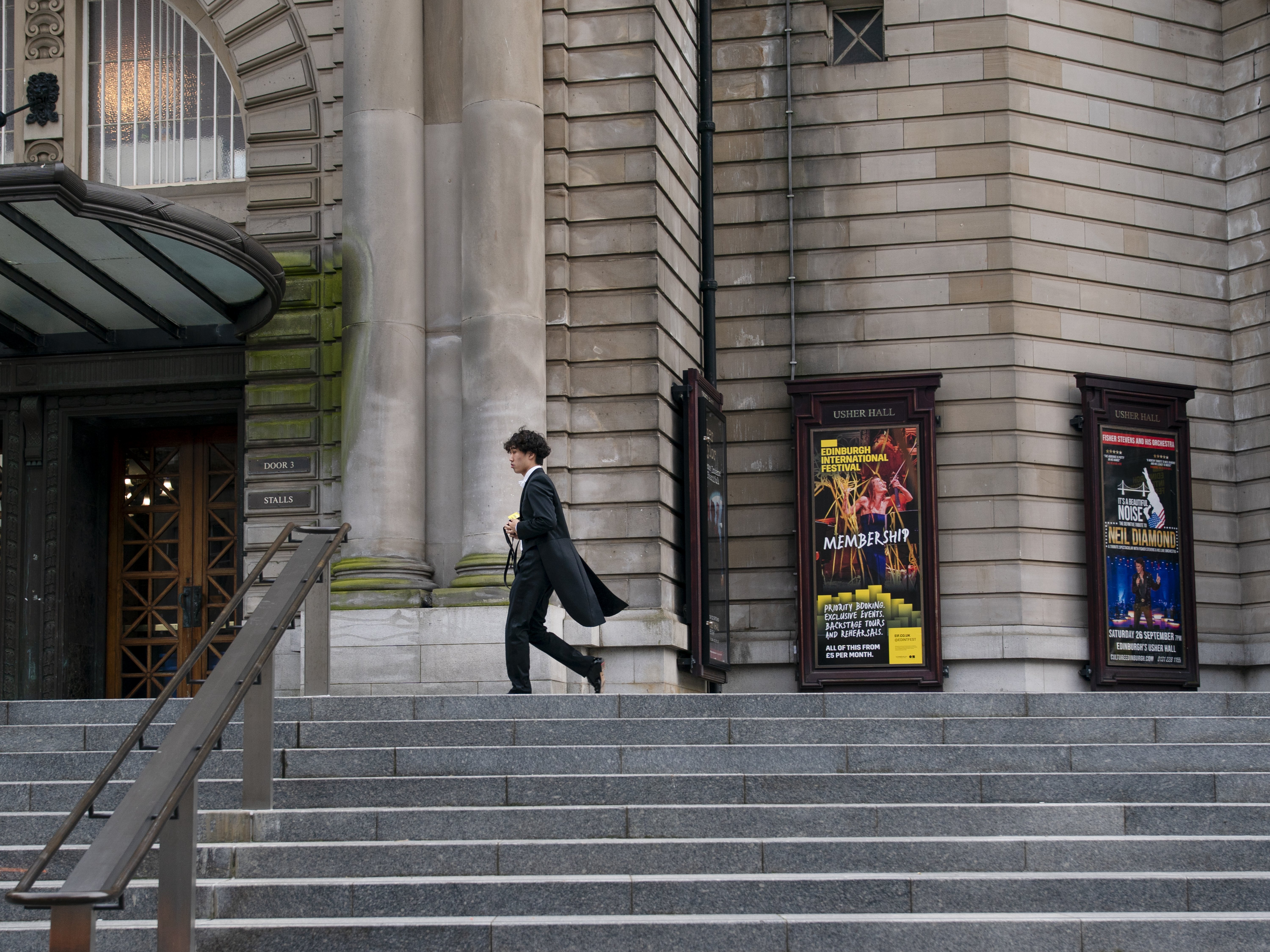 A person in dark clothing walks up wide stone steps outside a historic building with posters for shows on the wall and metal railings along the stairs.
