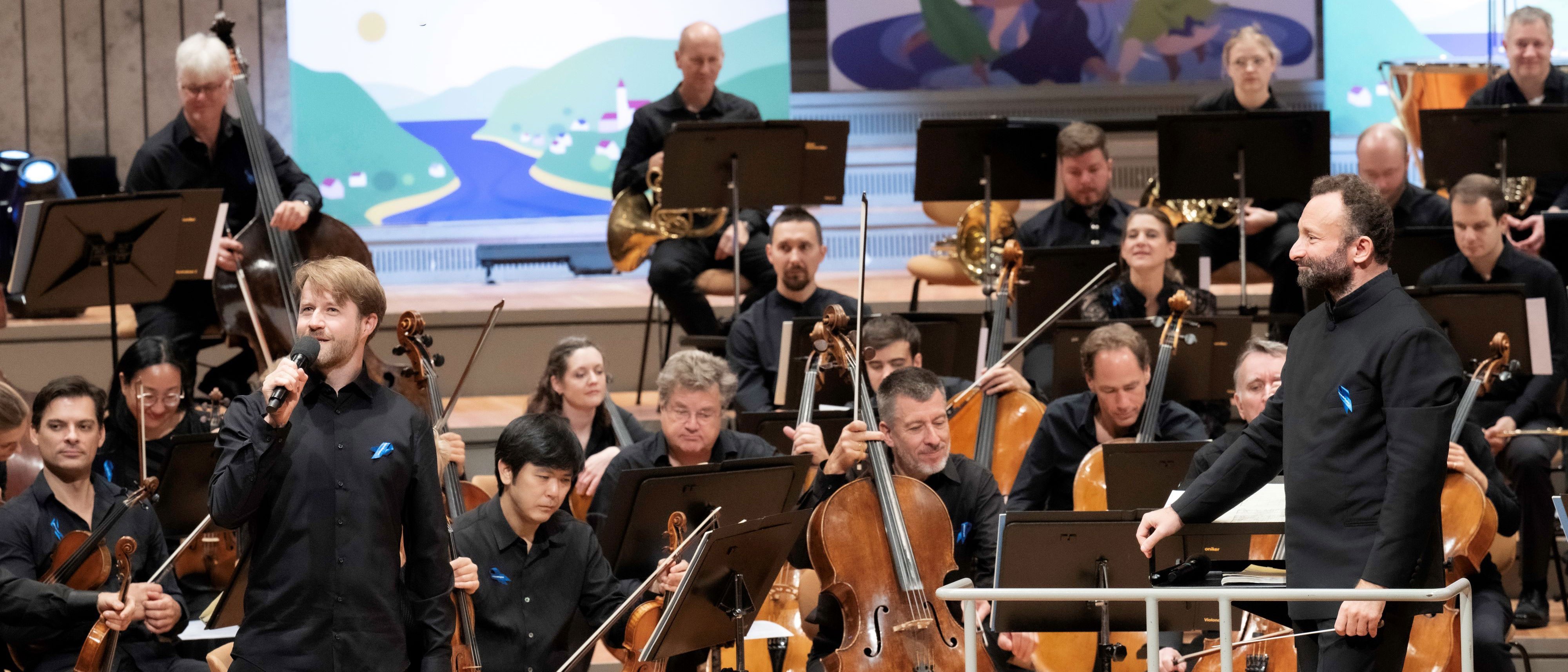 Auf der Bühne sind Orchestermitglieder mit Kirill Petrenko, mehreren Cellisten und anderen Musiker, die verschiedene Instrumente spielen. Im Hintergrund sind zwei große Kunstwerke zu sehen.