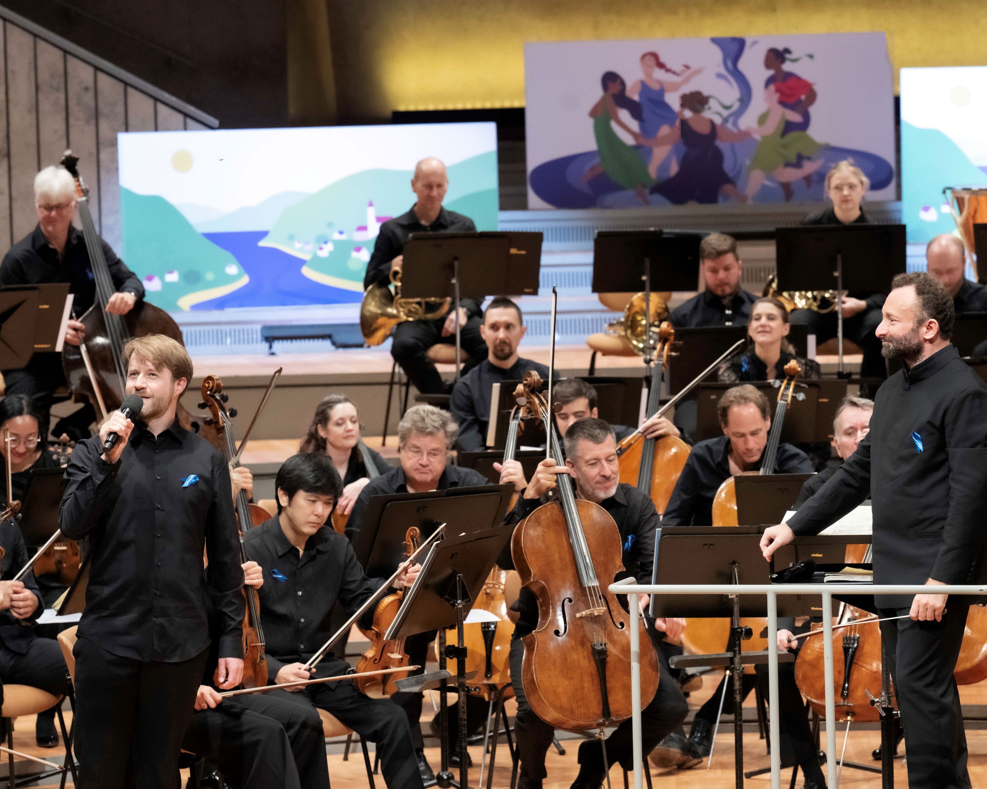 Auf der Bühne sind Orchestermitglieder mit Kirill Petrenko, mehreren Cellisten und anderen Musiker, die verschiedene Instrumente spielen. Im Hintergrund sind zwei große Kunstwerke zu sehen.