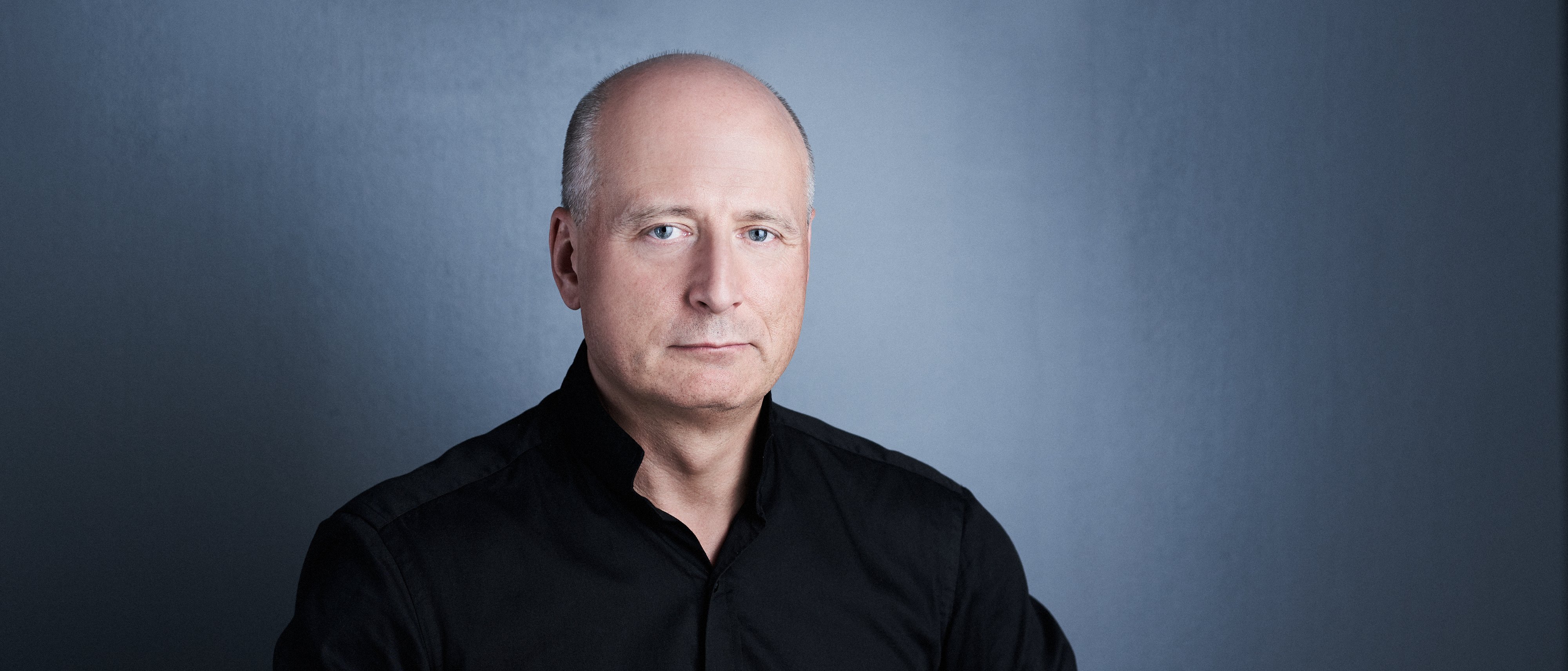 Ein Mann mit einer Halbglatze und in einem schwarzen Hemd sitzt vor einem neutralen Hintergrund. Er schaut neutral in die Kamera.