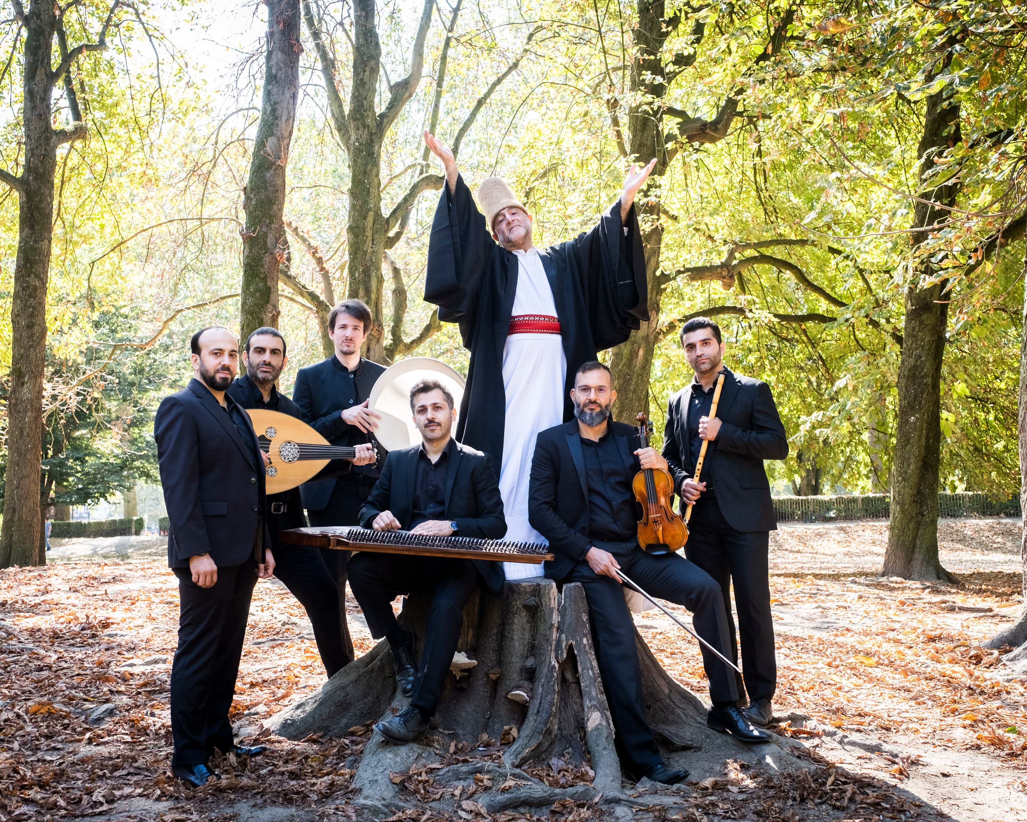 Sieben Männer in formeller schwarzer Kleidung posieren in einem sonnenbeschienenen Wald; einer von ihnen, gekleidet in Gewand und Hut, steht mit erhobenen Armen auf einem Baumstumpf, während andere traditionelle Musikinstrumente um ihn herum halten.