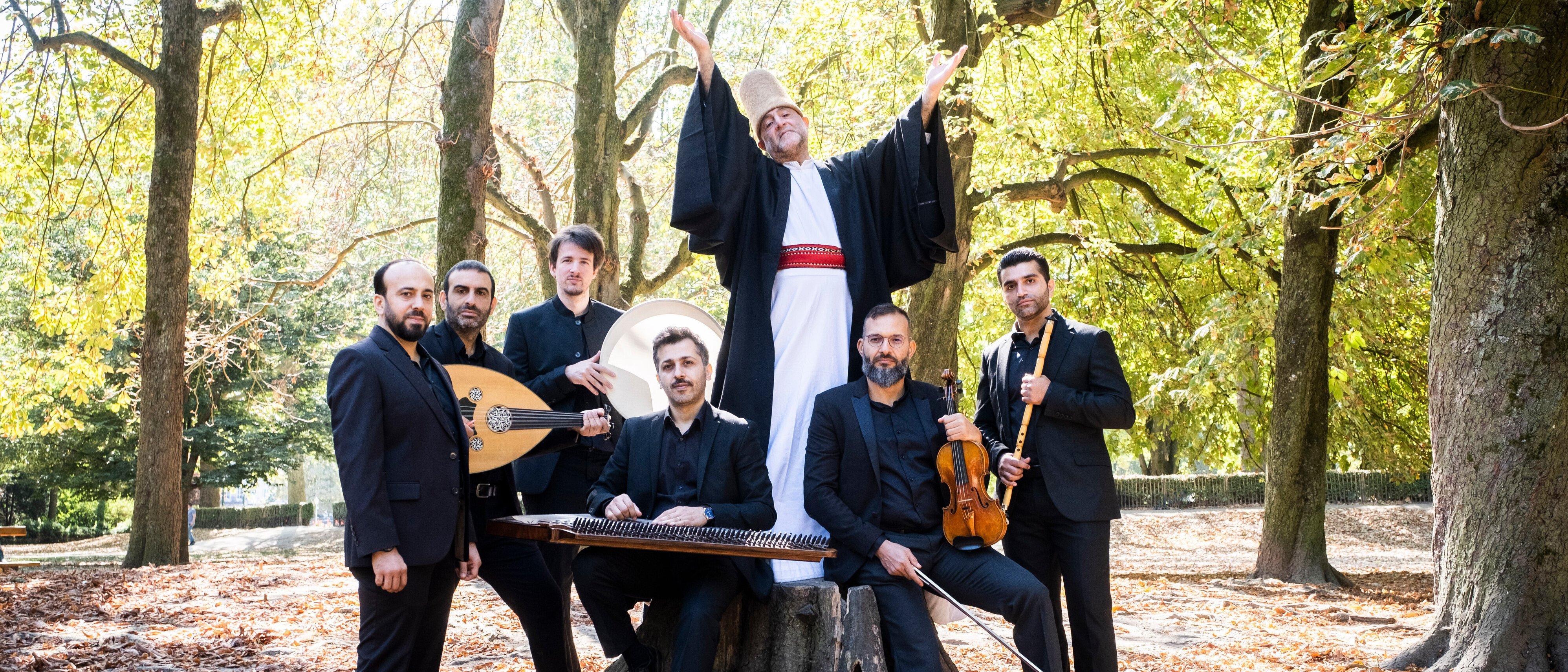 Sieben Männer in formeller schwarzer Kleidung posieren in einem sonnenbeschienenen Wald; einer von ihnen, gekleidet in Gewand und Hut, steht mit erhobenen Armen auf einem Baumstumpf, während andere traditionelle Musikinstrumente um ihn herum halten.