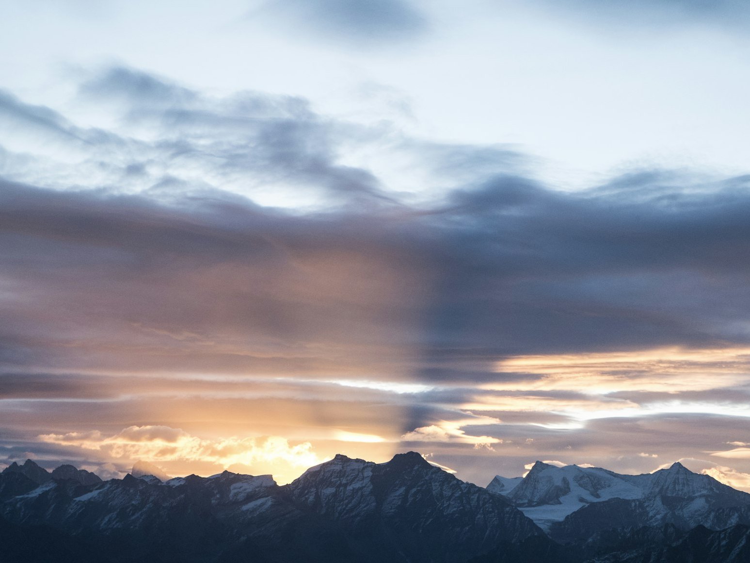 Verschneite Berggipfel unter einem dramatischen Himmel, an dem bei Sonnenaufgang oder Sonnenuntergang Sonnenstrahlen und Schatten durch bunte Wolken brechen.