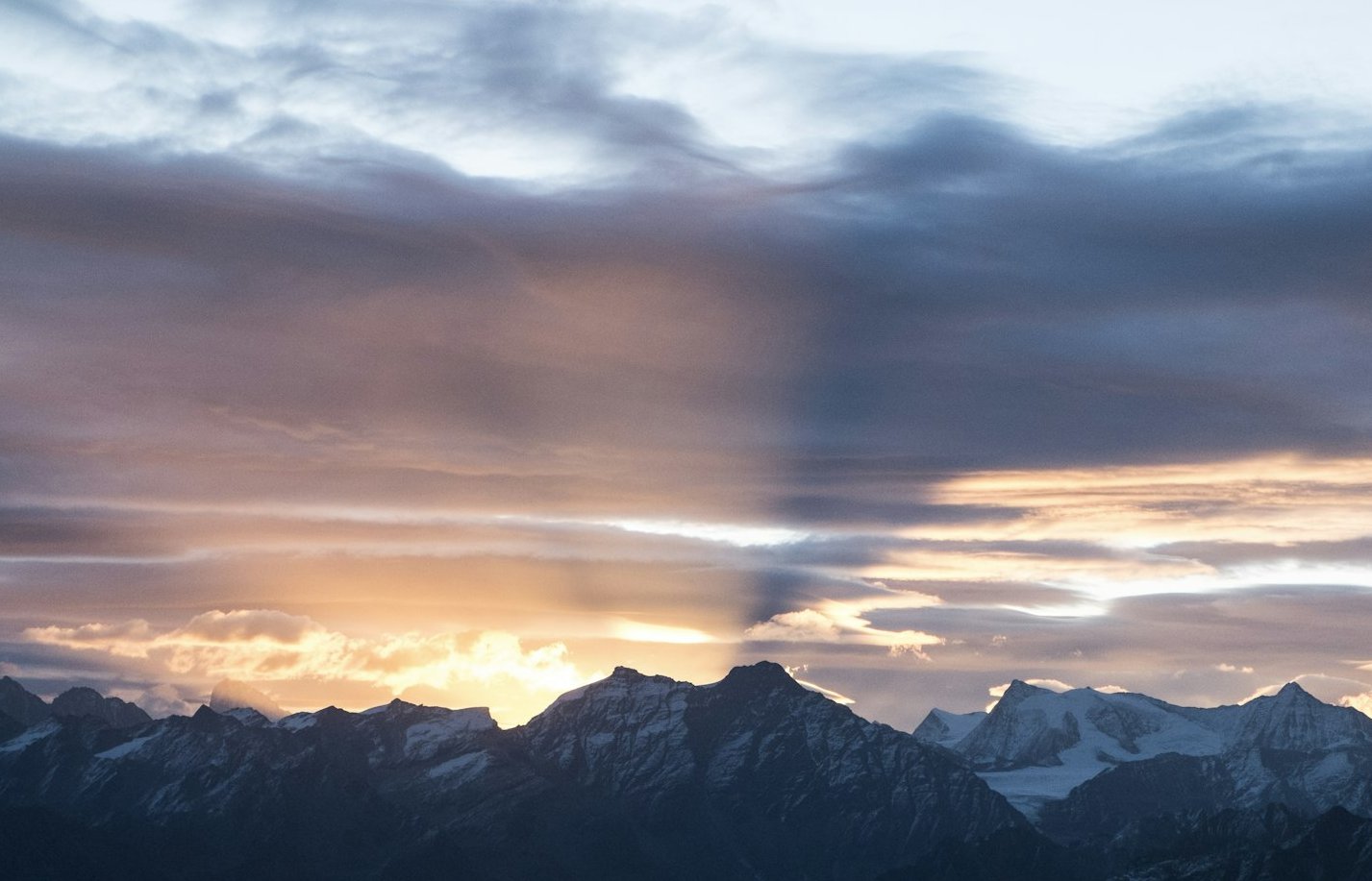 Verschneite Berggipfel unter einem dramatischen Himmel, an dem bei Sonnenaufgang oder Sonnenuntergang Sonnenstrahlen und Schatten durch bunte Wolken brechen.