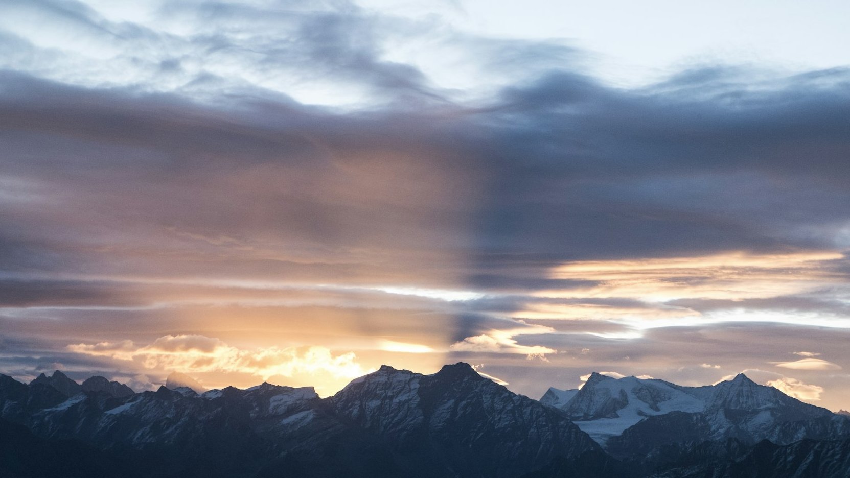 Sunset over snow-capped mountains, with rays of sunlight streaming through dramatic clouds, casting shadows and warm light across the sky.