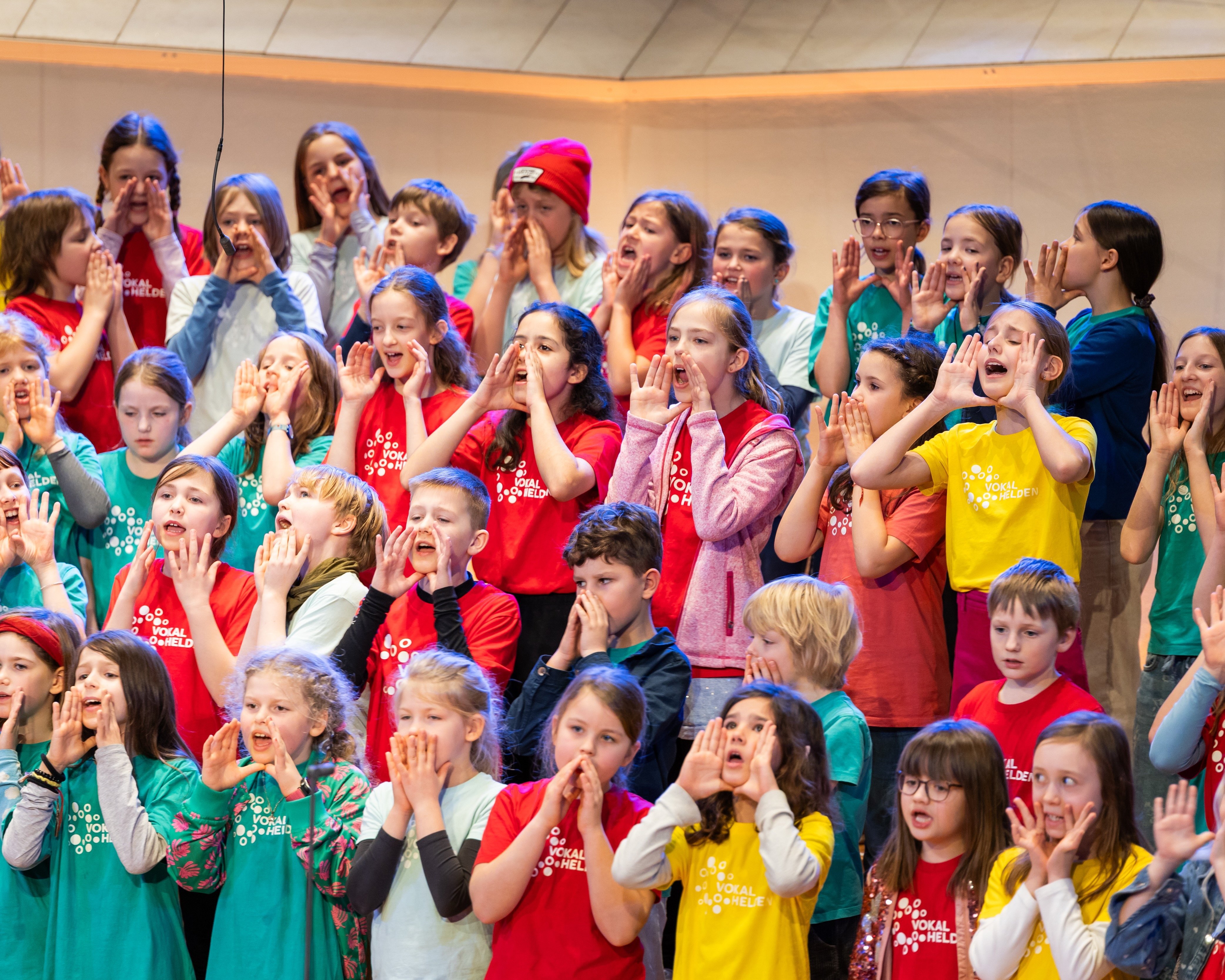 Eine große Gruppe von Kindern, die bunte Hemden tragen, steht in einem Chor auf den Rängen. Viele halten sich die Hände vor den Mund, während sie bei einer Aufführung oder Probe begeistert singen oder schreien.
