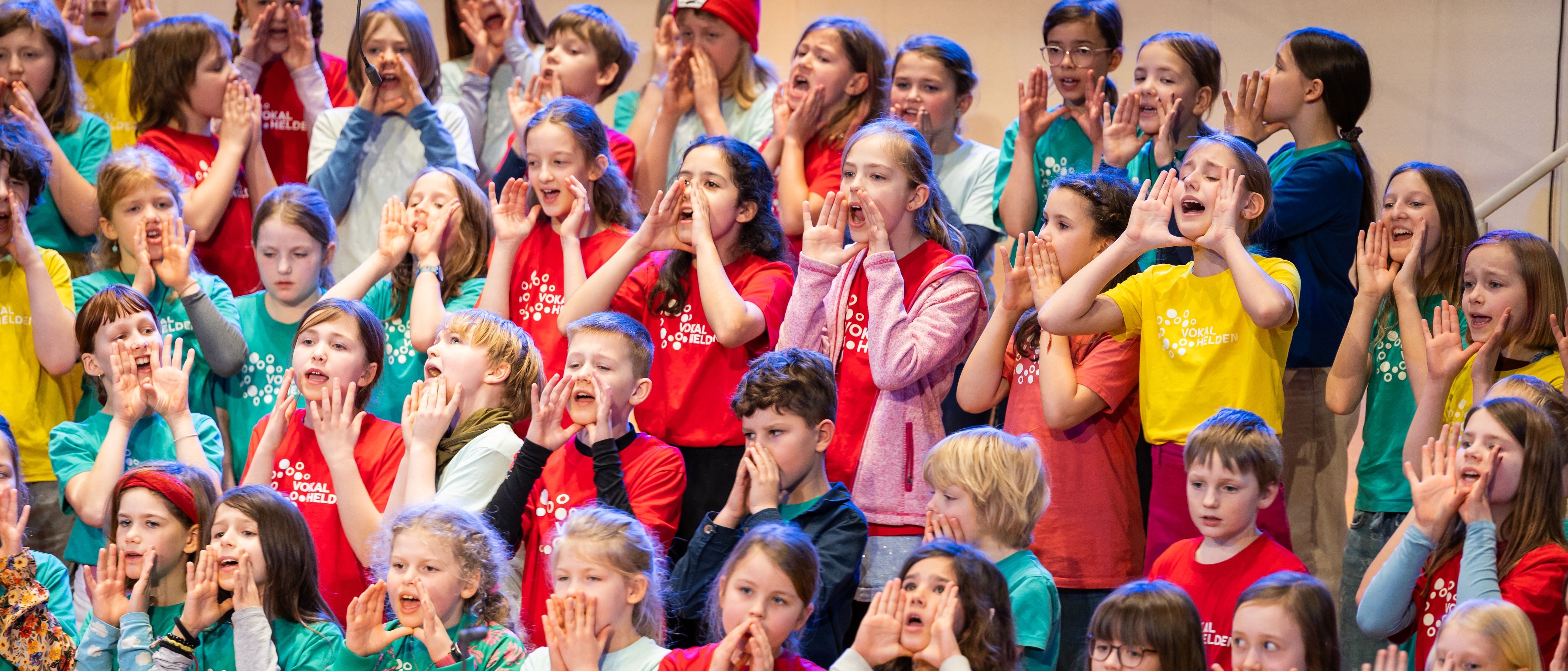 Eine große Gruppe von Kindern, die bunte Hemden tragen, steht in einem Chor auf den Rängen. Viele halten sich die Hände vor den Mund, während sie bei einer Aufführung oder Probe begeistert singen oder schreien.