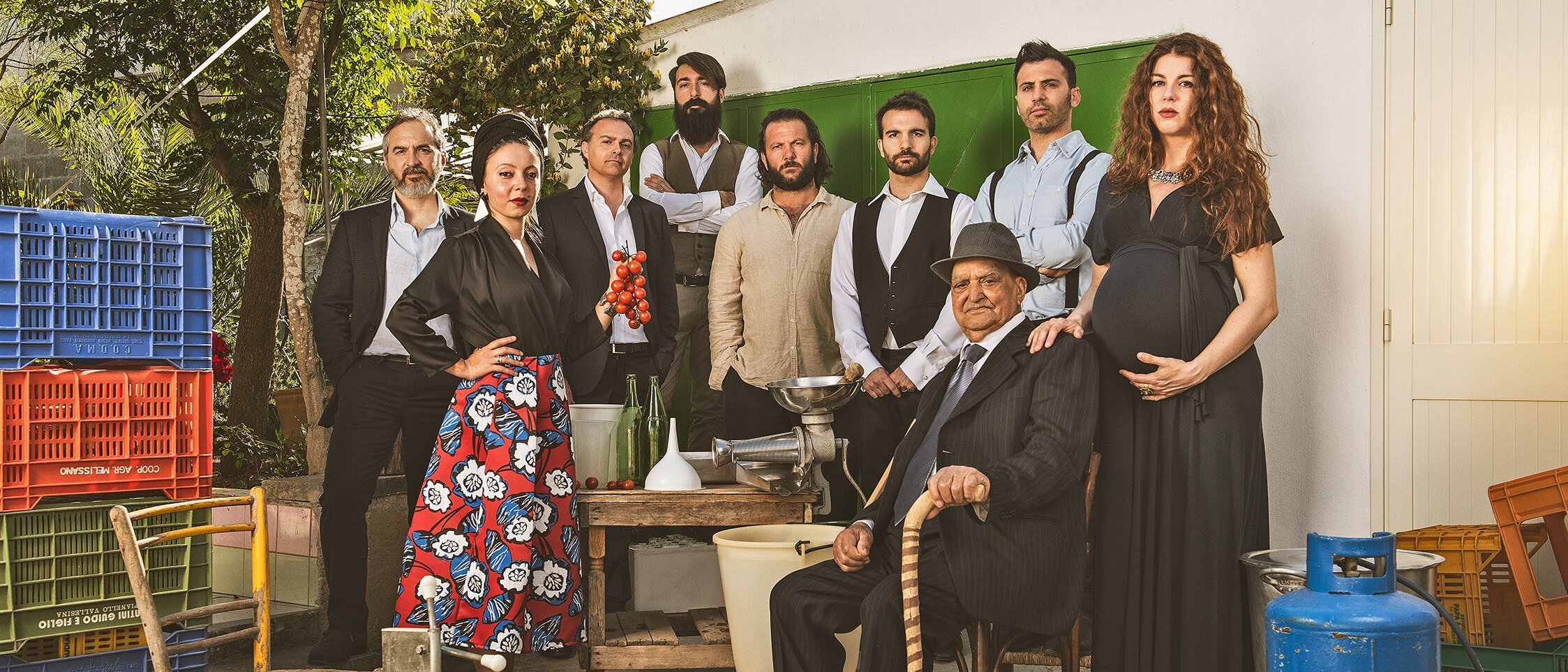 Neun Erwachsene posieren gemeinsam im Freien mit Kisten, Flaschen, Schüsseln und einem blauen Benzintank. Eine Frau ist schwanger, ein Mann sitzt mit einem Stock und die anderen stehen in verschiedenen Posen und sind halb-formal gekleidet.