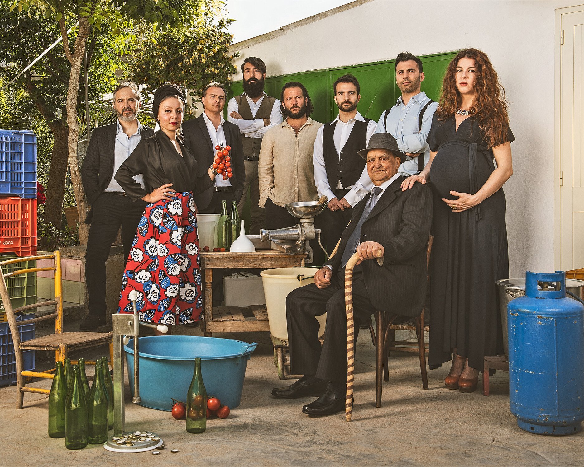 Neun Erwachsene posieren gemeinsam im Freien mit Kisten, Flaschen, Schüsseln und einem blauen Benzintank. Eine Frau ist schwanger, ein Mann sitzt mit einem Stock und die anderen stehen in verschiedenen Posen und sind halb-formal gekleidet.